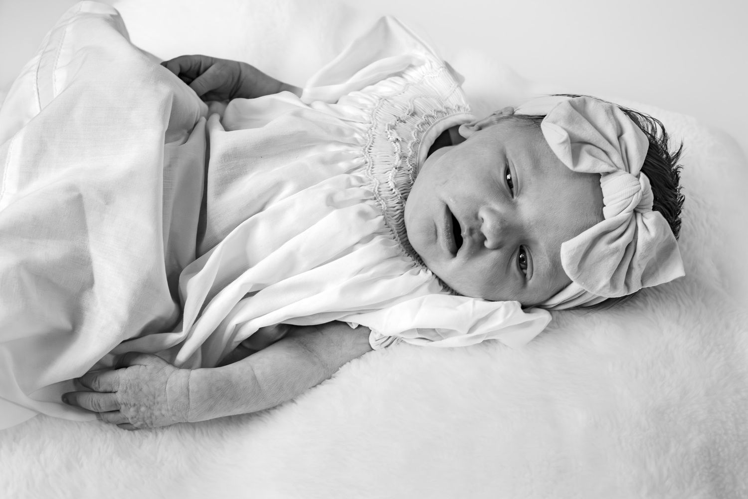 Black and white portrait of a newborn baby girl wearing a white bow headband, peacefully resting on a soft blanket.