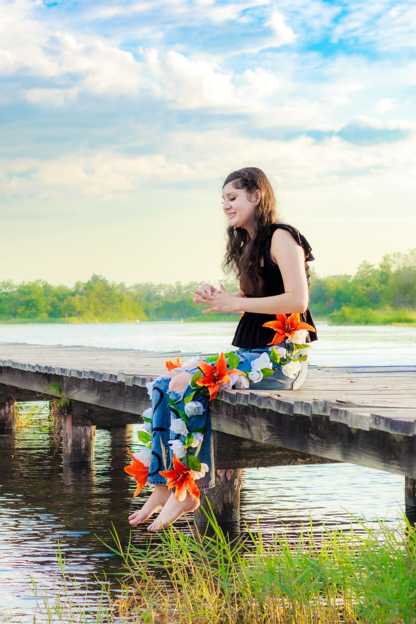 Young woman sitting on wooden dock over lake, wearing floral jeans, smiling, with orange lilies, green trees background.