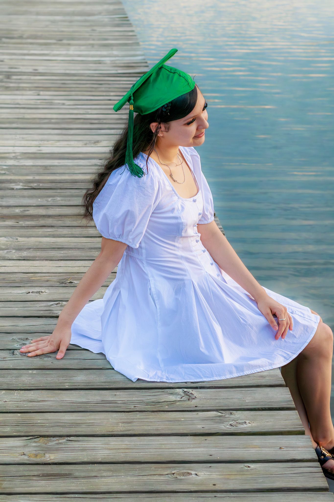 Young woman in white dress and green graduation cap sitting on wooden dock by calm water.