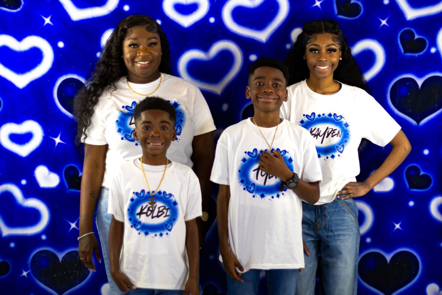 Four kids smiling in matching white logo t-shirts against a blue heart backdrop.