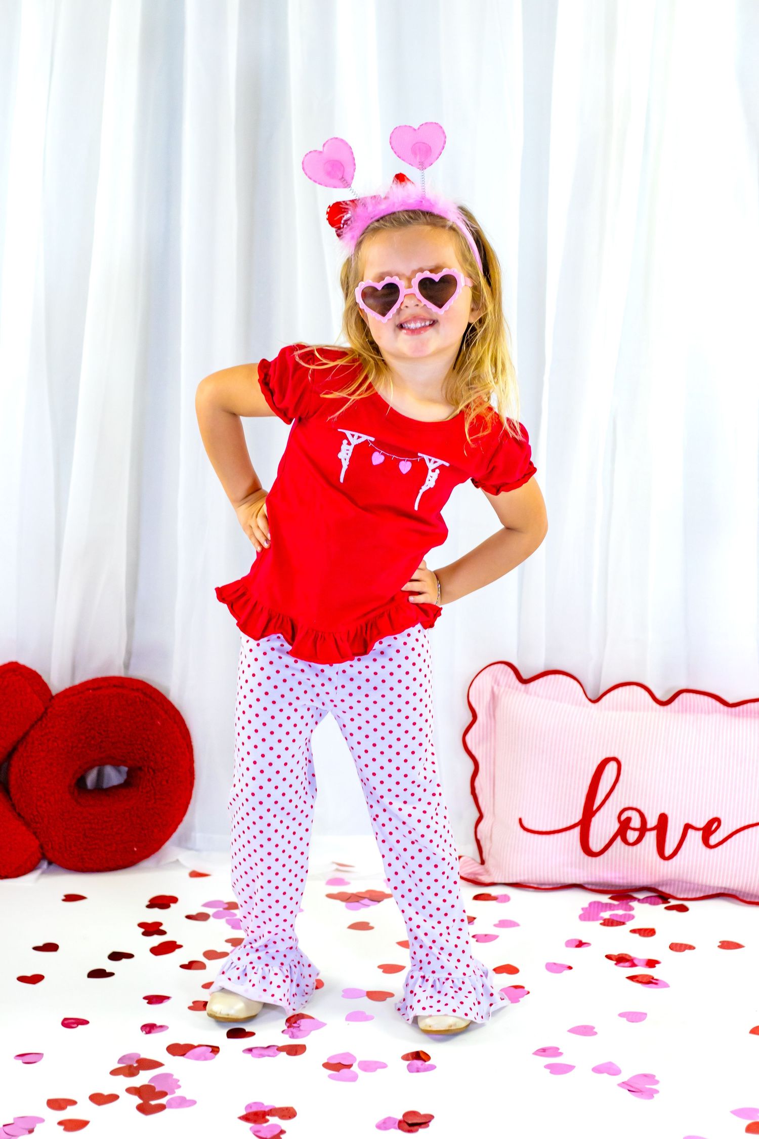 Young girl in red Valentine's outfit with heart sunglasses posing among confetti and love pillow.