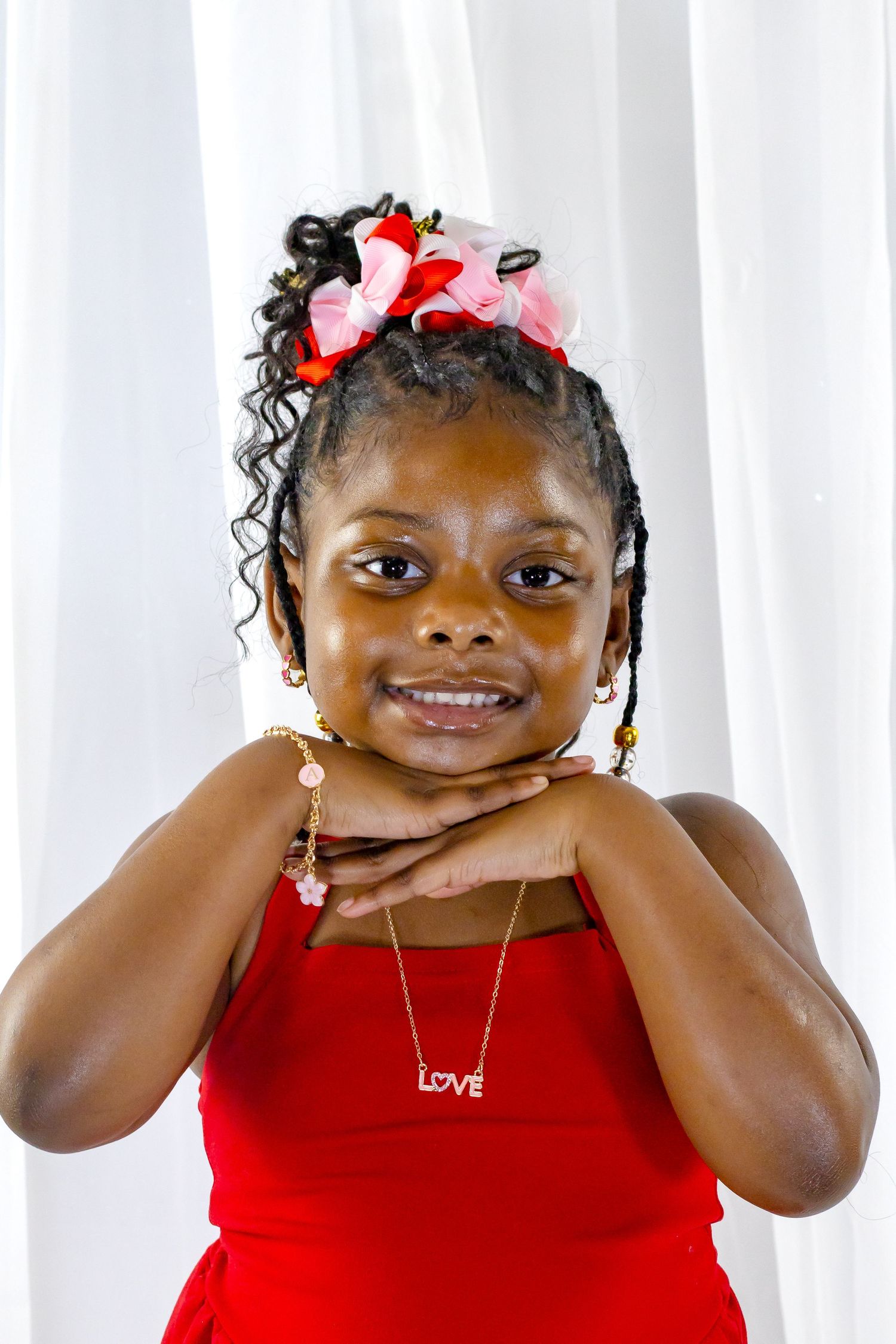 Smiling young Black girl in red dress wearing a Love necklace with hands under chin.