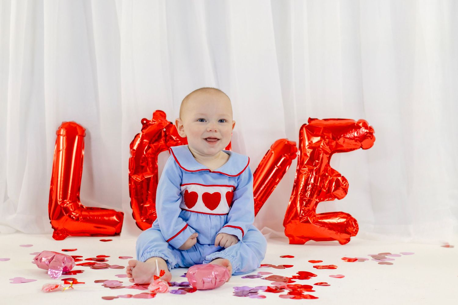 Smiling baby in blue heart romper sits before a red LOVE balloon with scattered heart confetti.