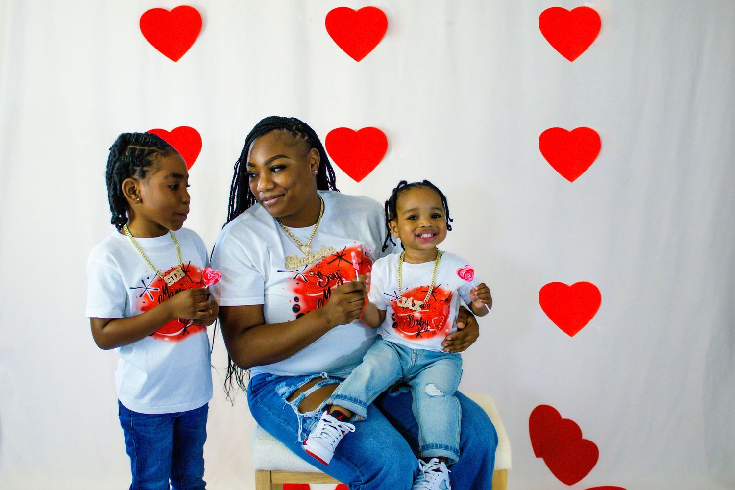 Mother and two children in matching shirts smile holding Valentine's gifts in front of heart backdrop.