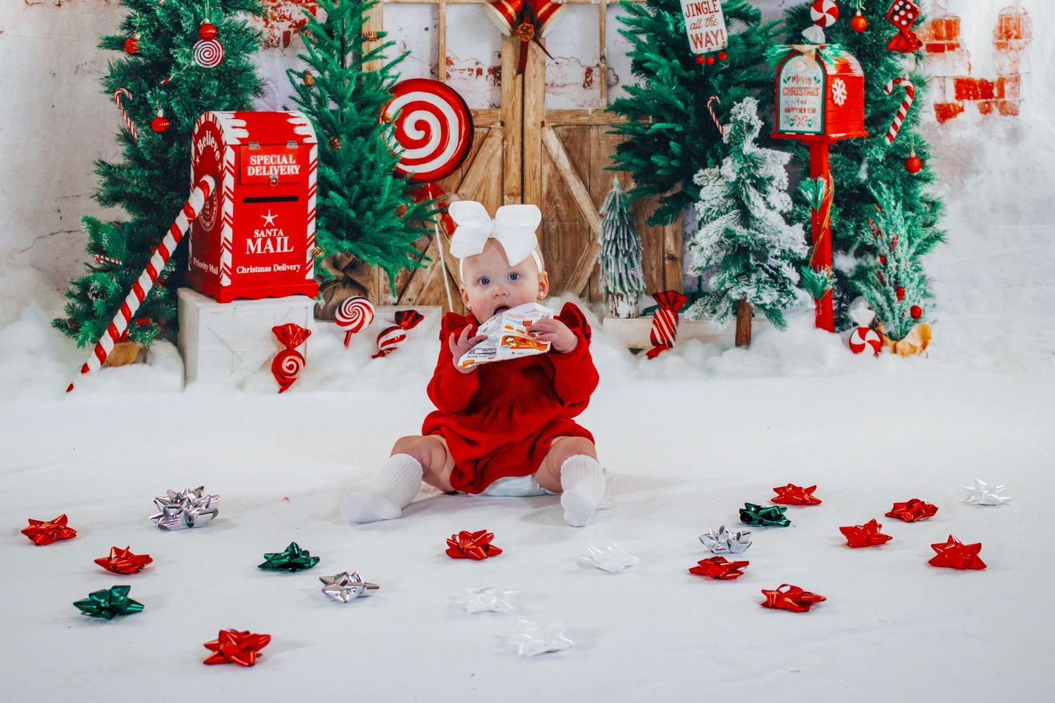 Baby girl in red dress with white bow sitting among scattered bows in a Christmas candy cane photo studio.