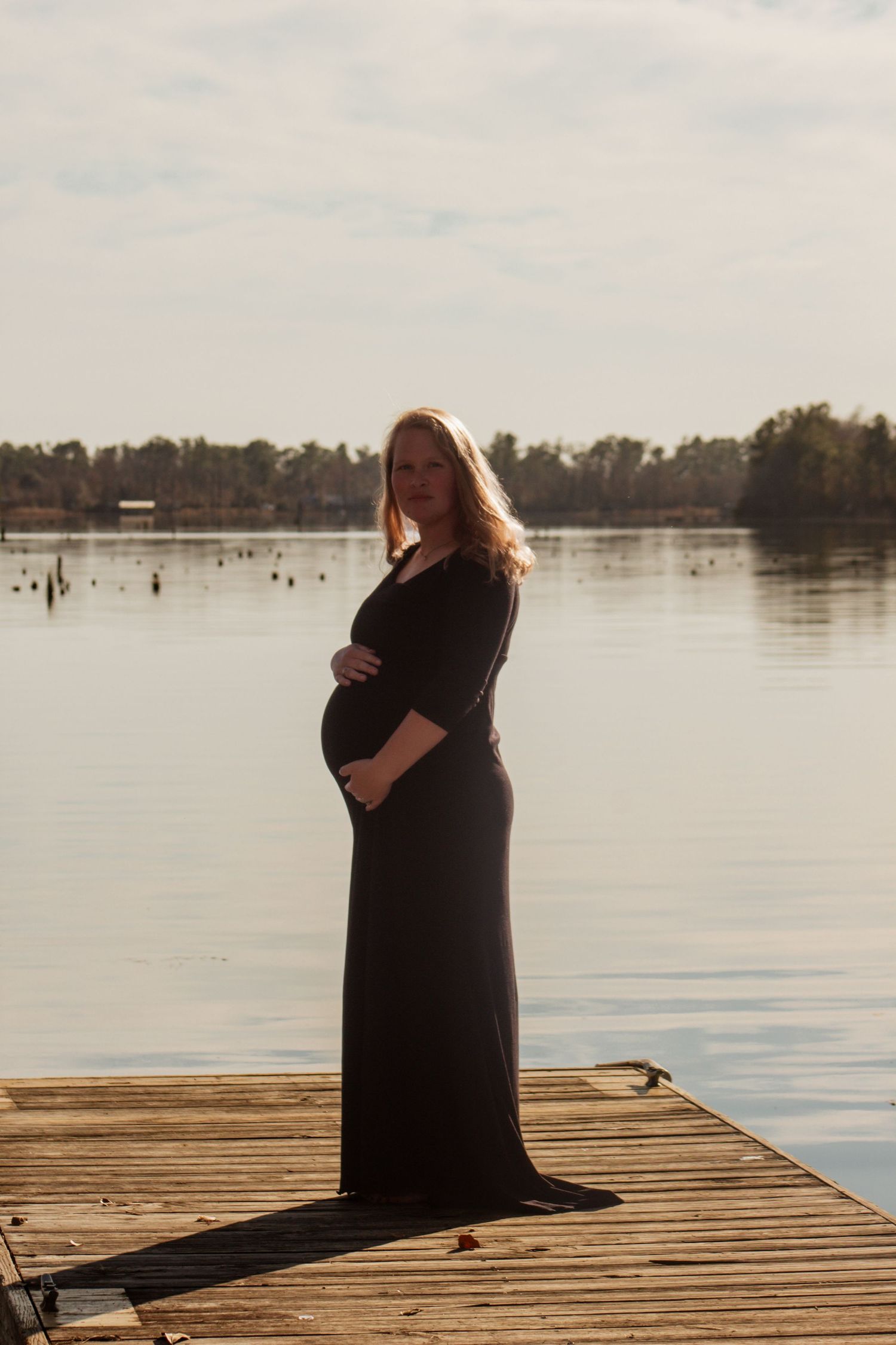 Pregnant woman in black maxi dress standing on wooden dock by calm lake holding baby bump at sunset.