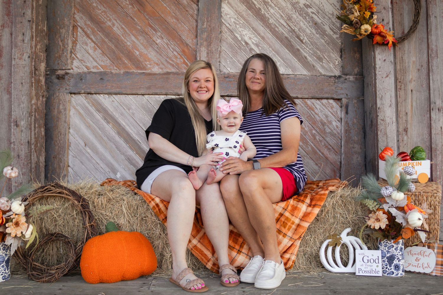 Two women and baby girl posing together on hay bales with autumn pumpkin decorations.