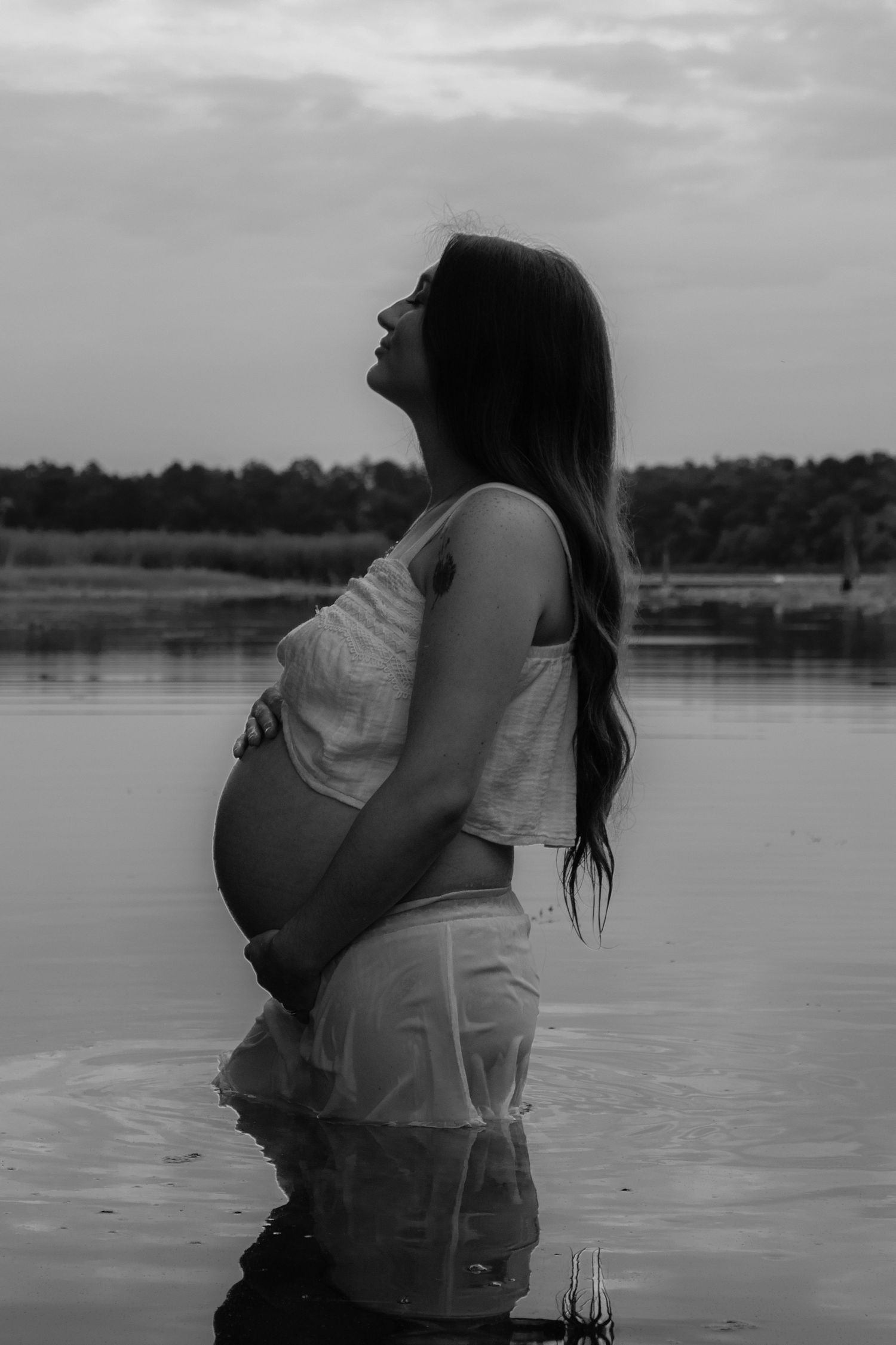 Black and white side profile of pregnant woman cradling baby bump while standing in lake water at sunset.