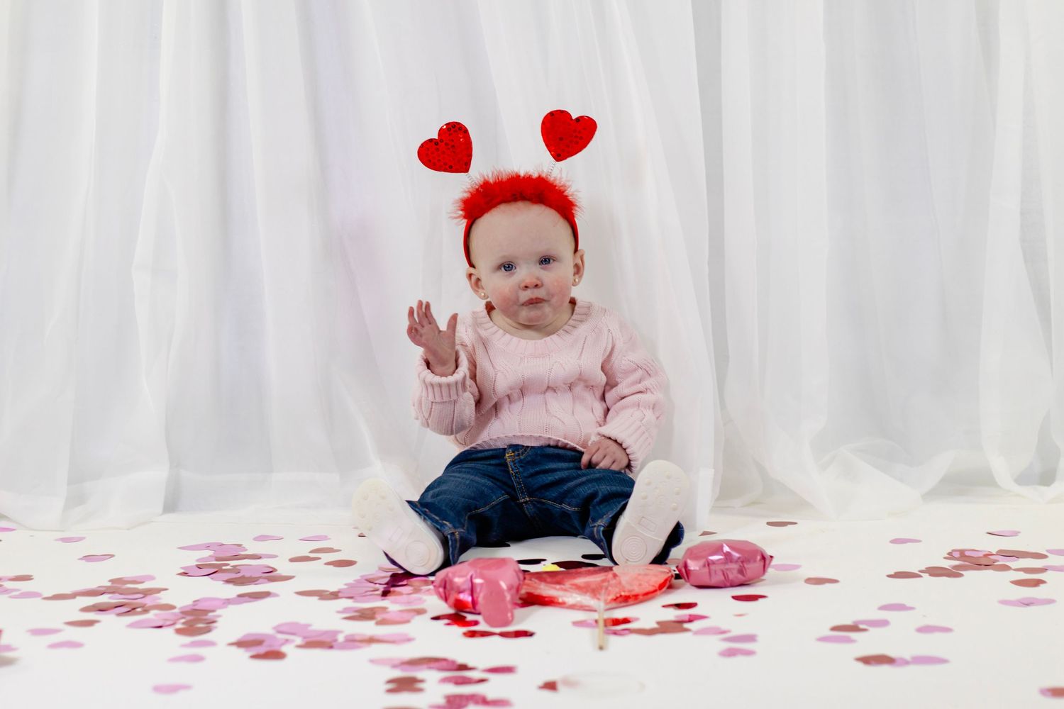 Sweet baby wearing red heart headband sitting among pink and red Valentine's Day heart confetti decorations.