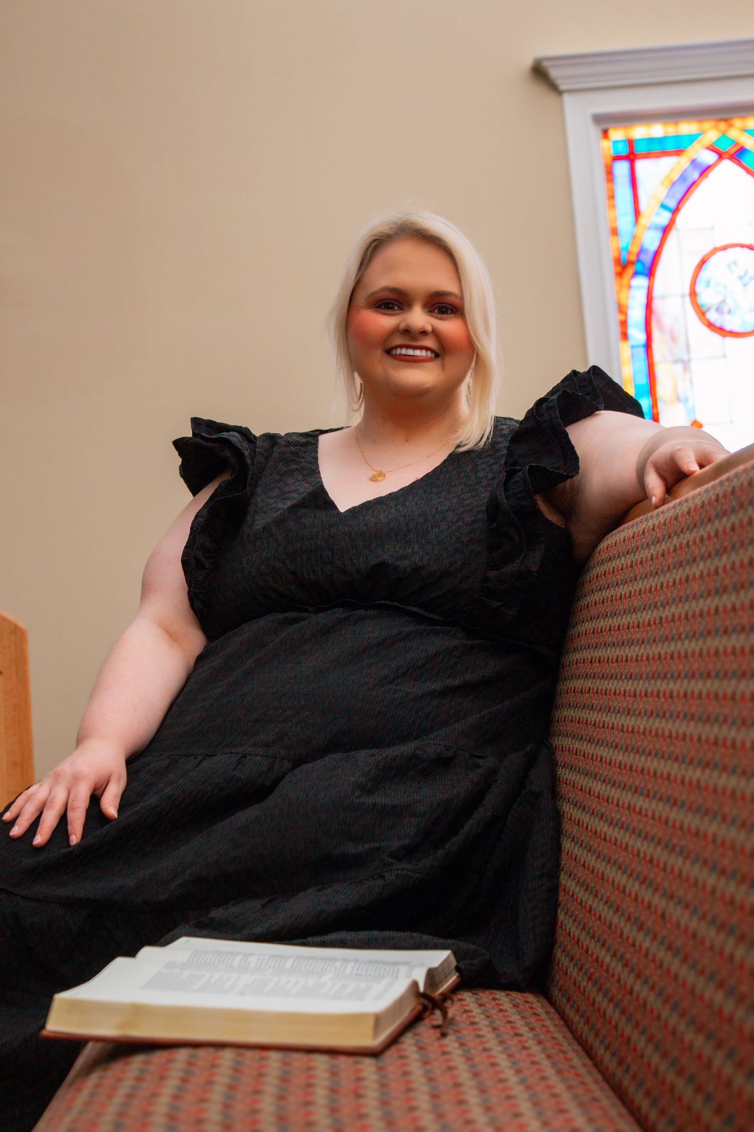 Smiling blonde woman in black dress relaxing on church pew with Bible and stained glass window.