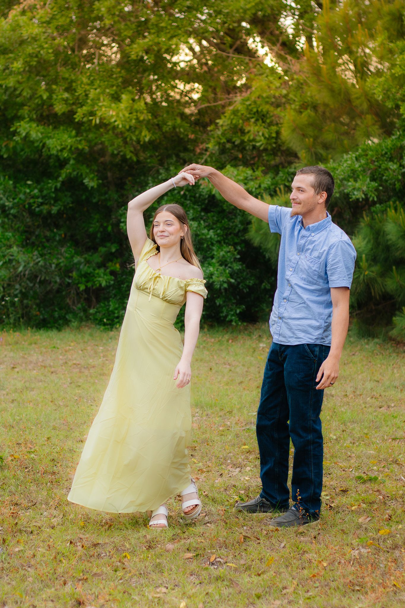 A young couple dances outdoors, woman in flowing yellow dress being twirled by man in blue shirt and jeans.