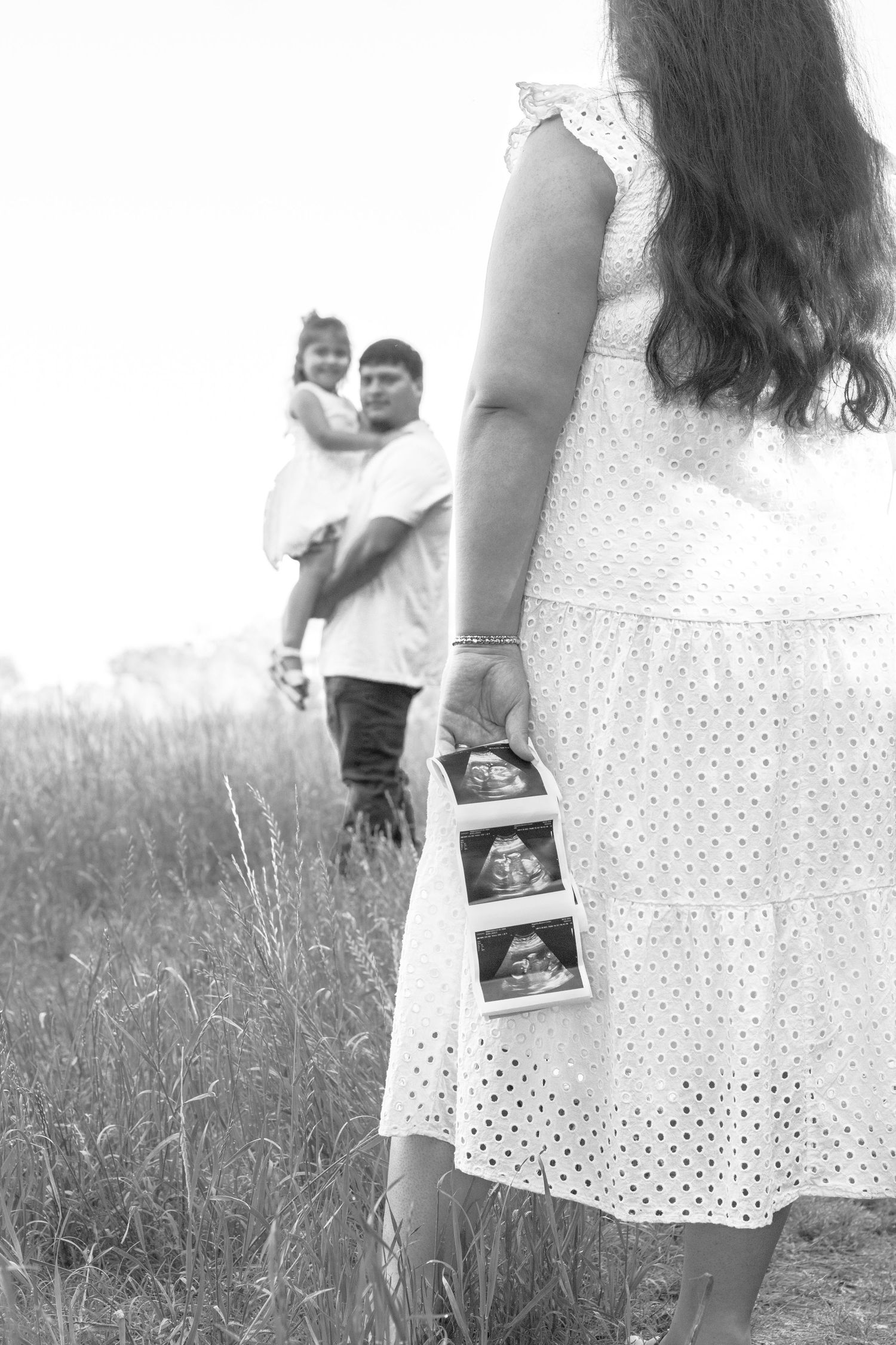 Black and white photo of pregnant woman holding ultrasound images in a field with family in background.