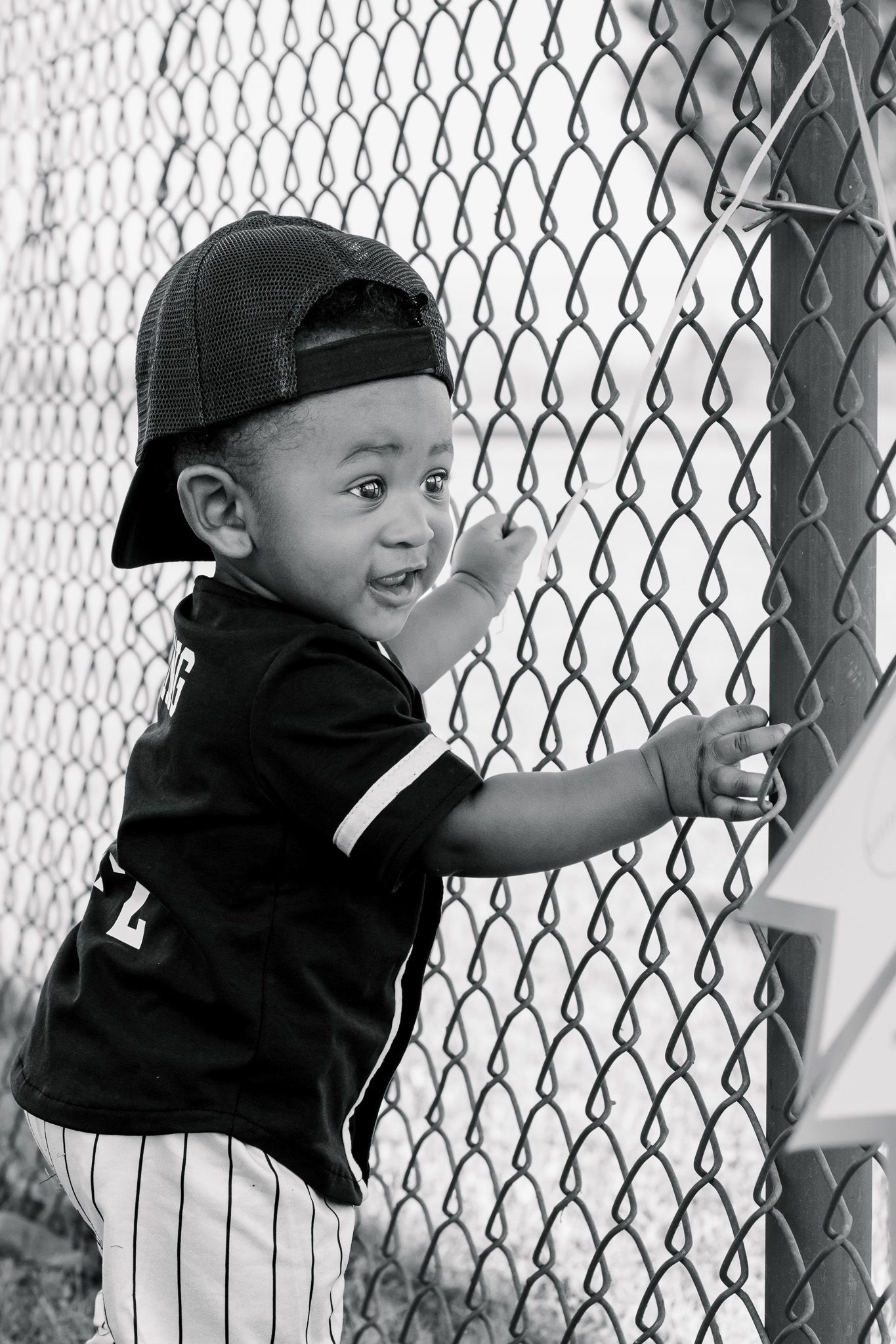 Black and white photo of a young boy in baseball uniform and backwards cap holding a chain-link fence.