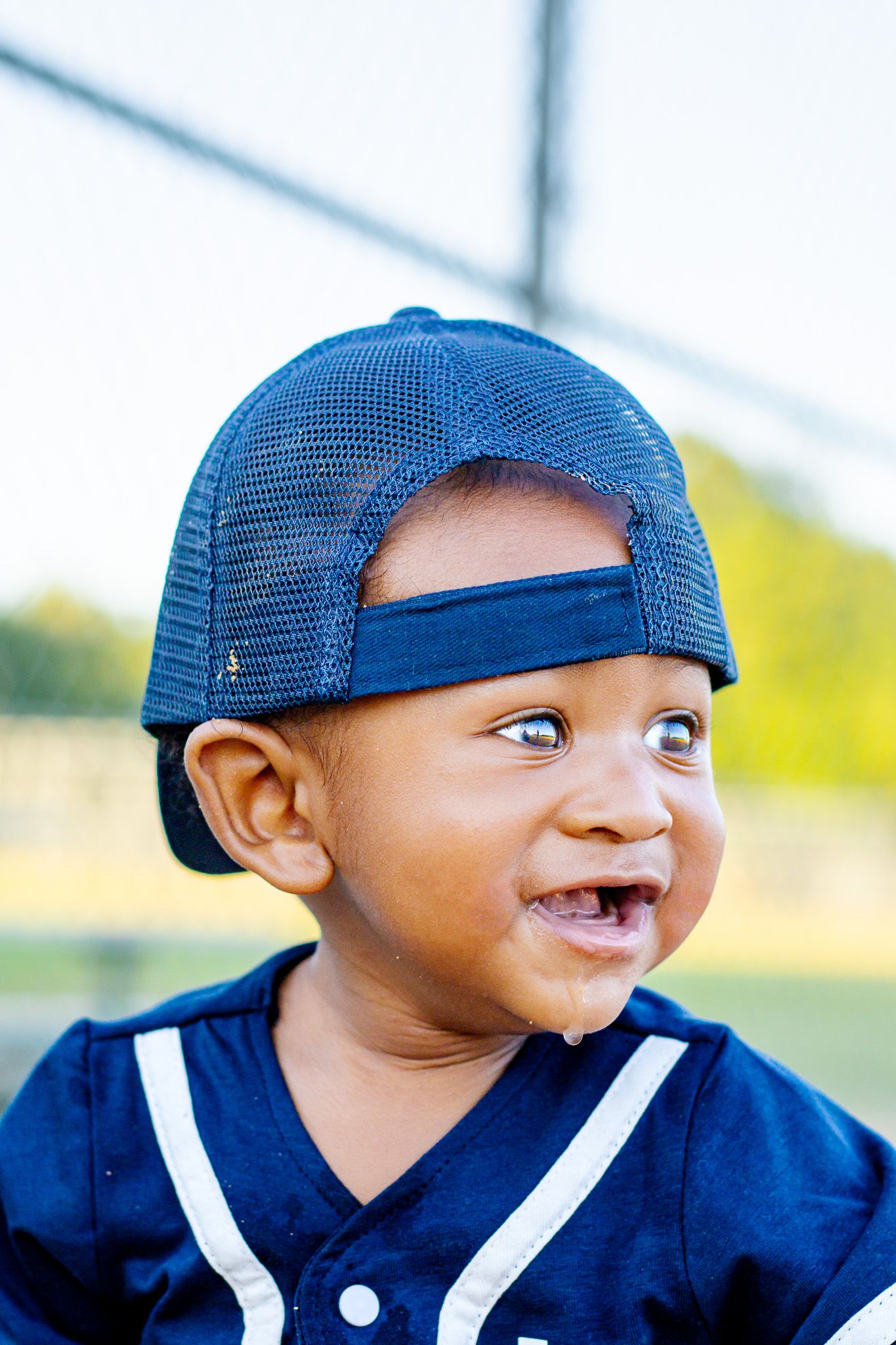 Smiling toddler boy wearing a backwards blue baseball cap and navy baseball uniform outdoors.