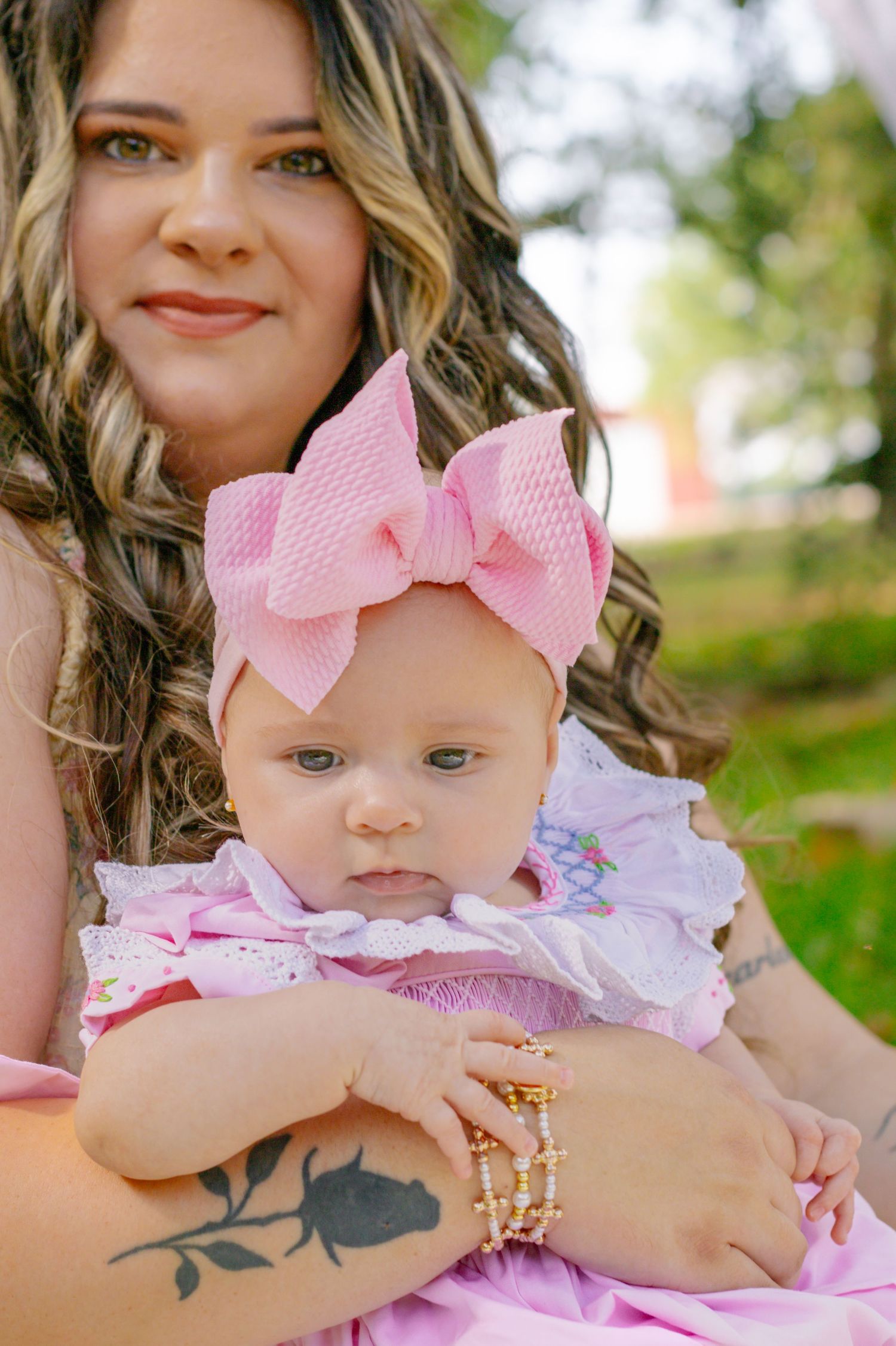 Close-up portrait of baby girl in pink smocked outfit with bow headband held by tattooed mother outdoors.