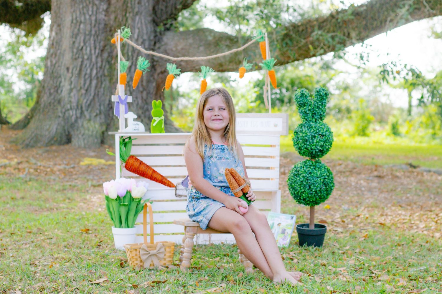 Girl in denim overalls sits on white bench with Easter carrot decorations and topiary outdoors.
