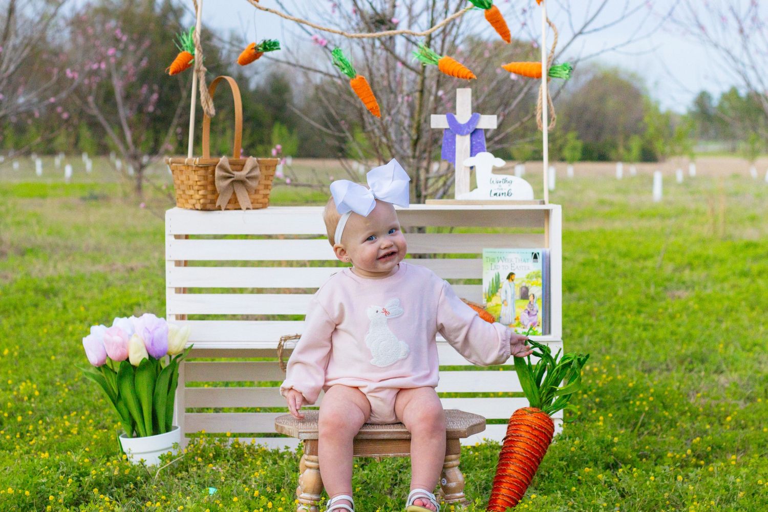 Cheerful baby in pink romper sits on bench with Easter decorations and tulips outdoors.