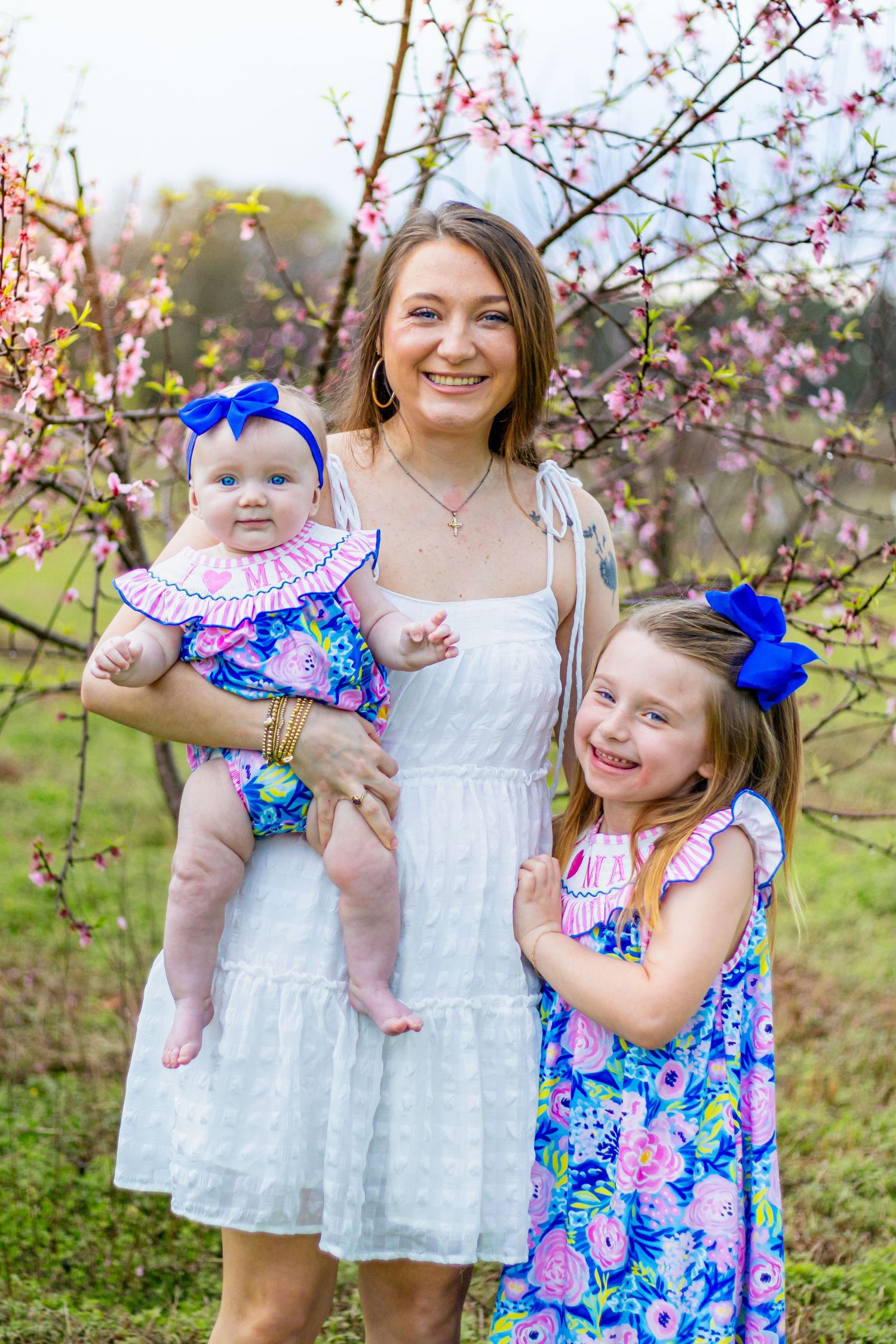 Smiling mother in white dress poses with two daughters in matching blue floral outfits by blossoms.