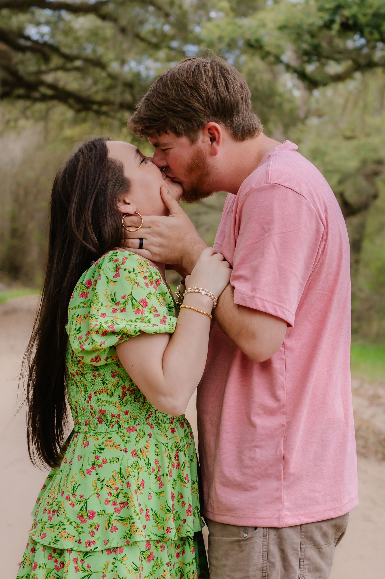 Couple sharing a romantic kiss on a dirt path with green trees in the background.