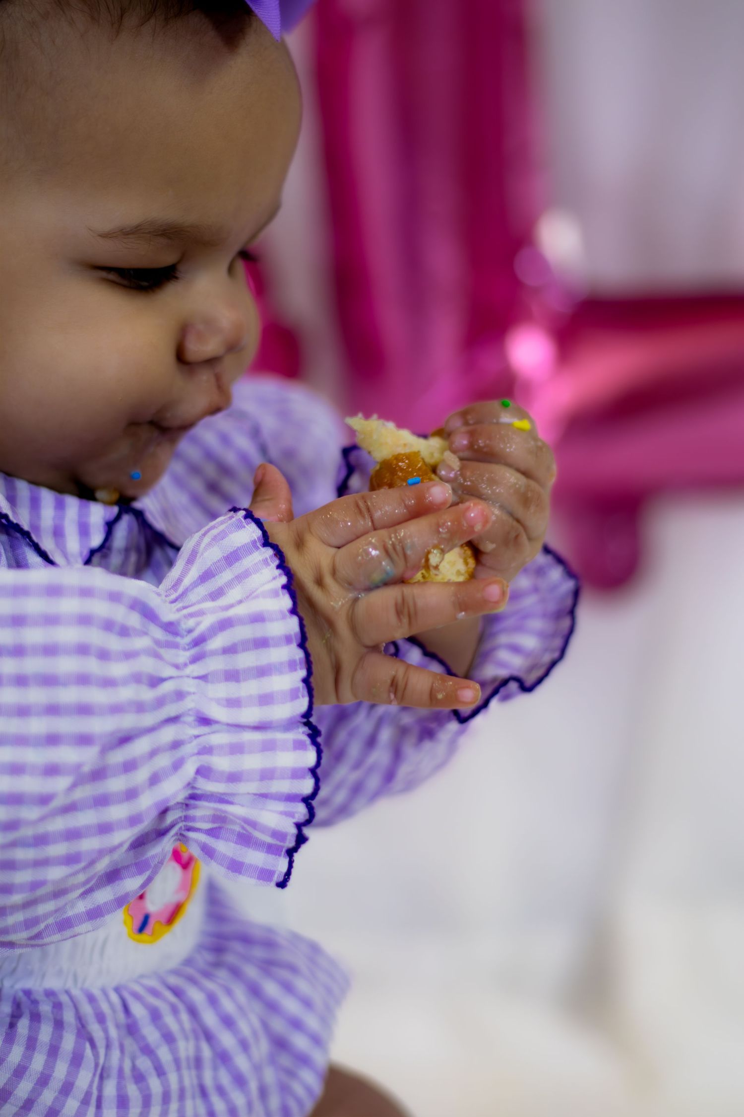 Close-up of baby girl with messy hands eating a donut in purple gingham outfit.