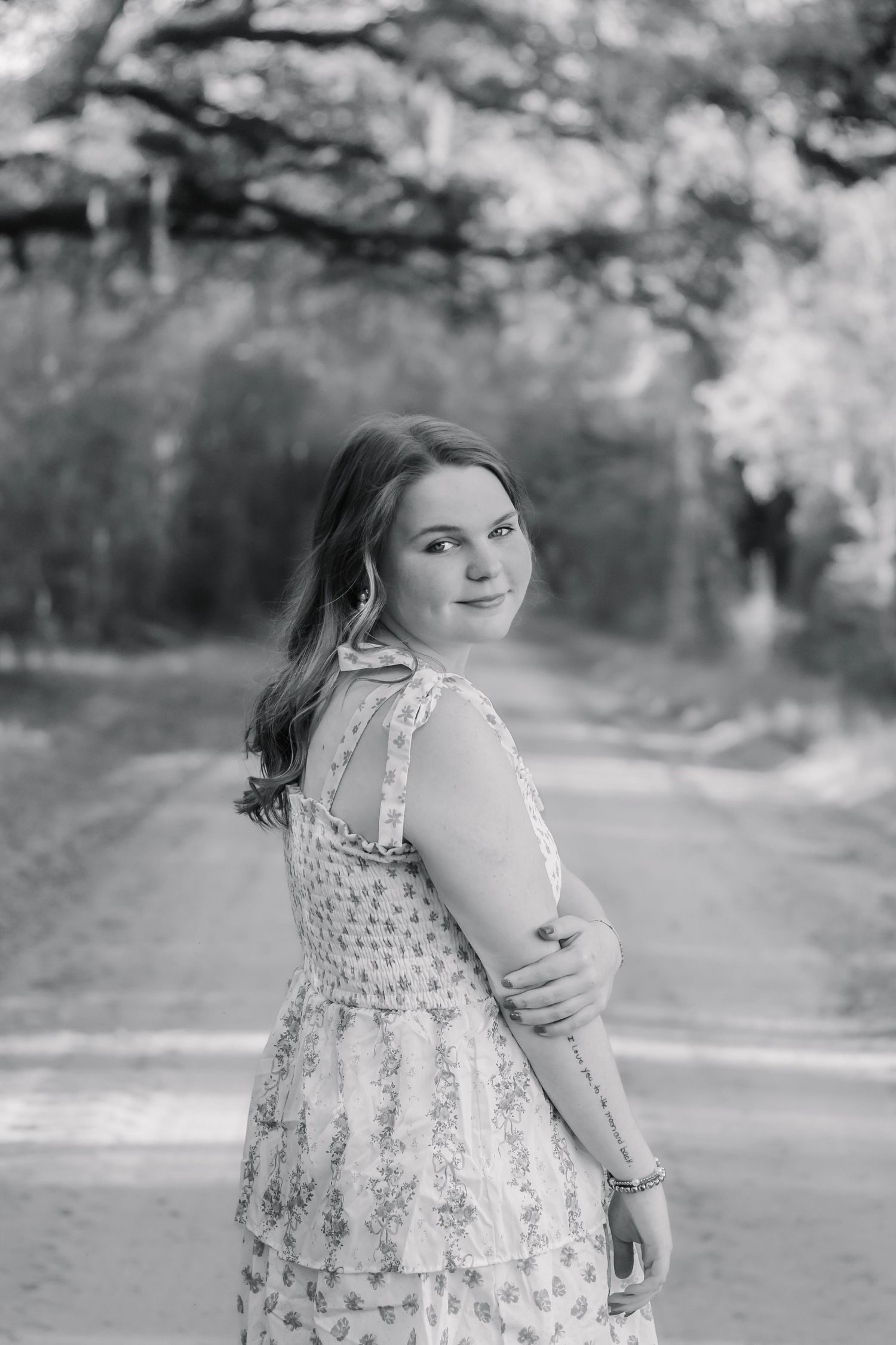 Black and white portrait of a smiling girl in a floral dress on a tree-lined dirt road.