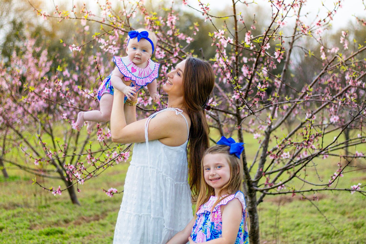 Mother lifting smiling baby girl while older daughter stands beside pink blossoming trees outdoors.