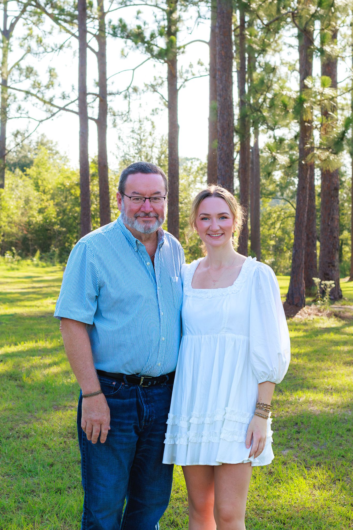 Smiling older man in blue shirt and young woman in white dress posing together outdoors among tall pine trees.