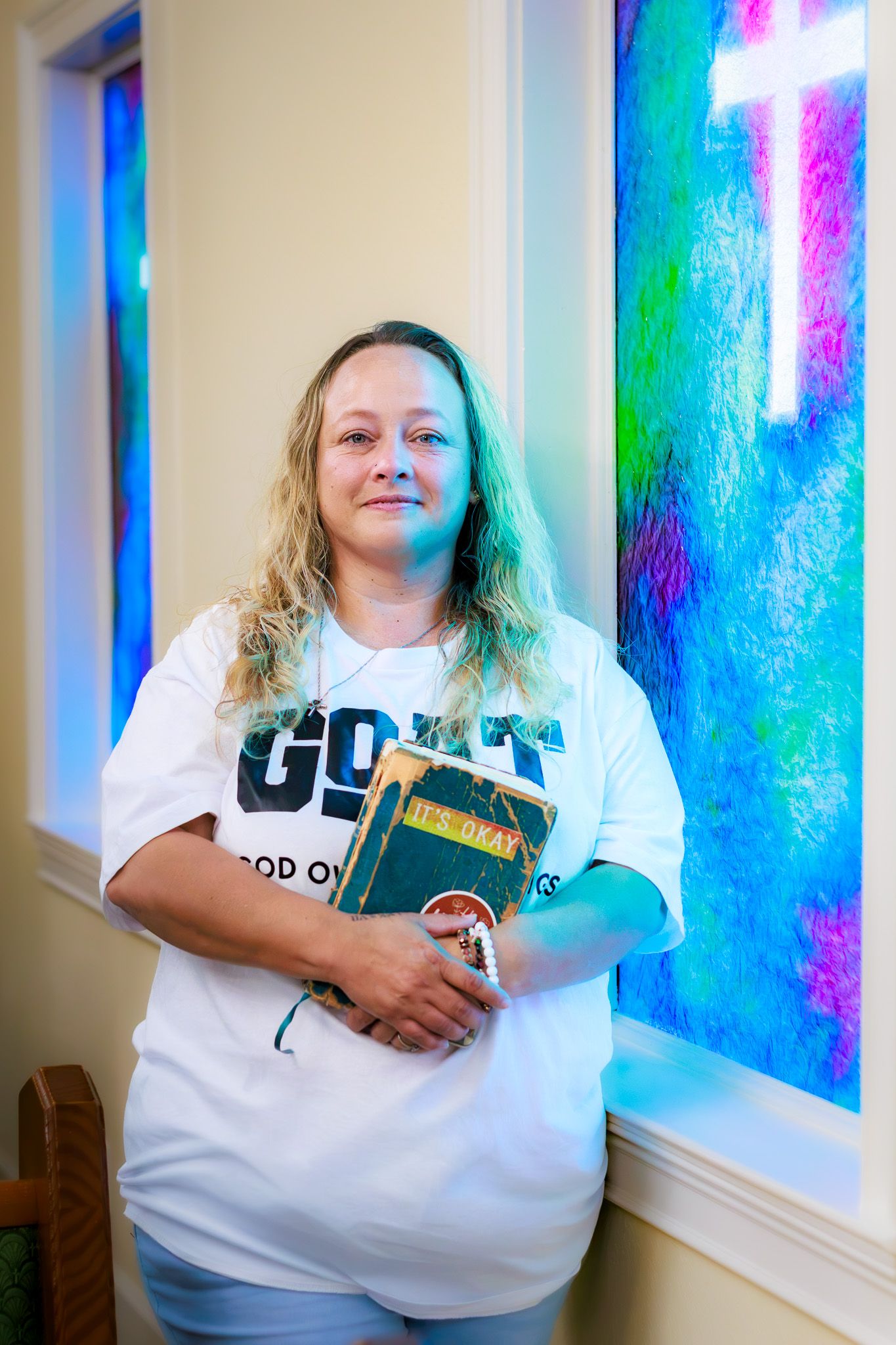 Woman holding a Bible stands before colorful stained glass windows with a cross in a church.