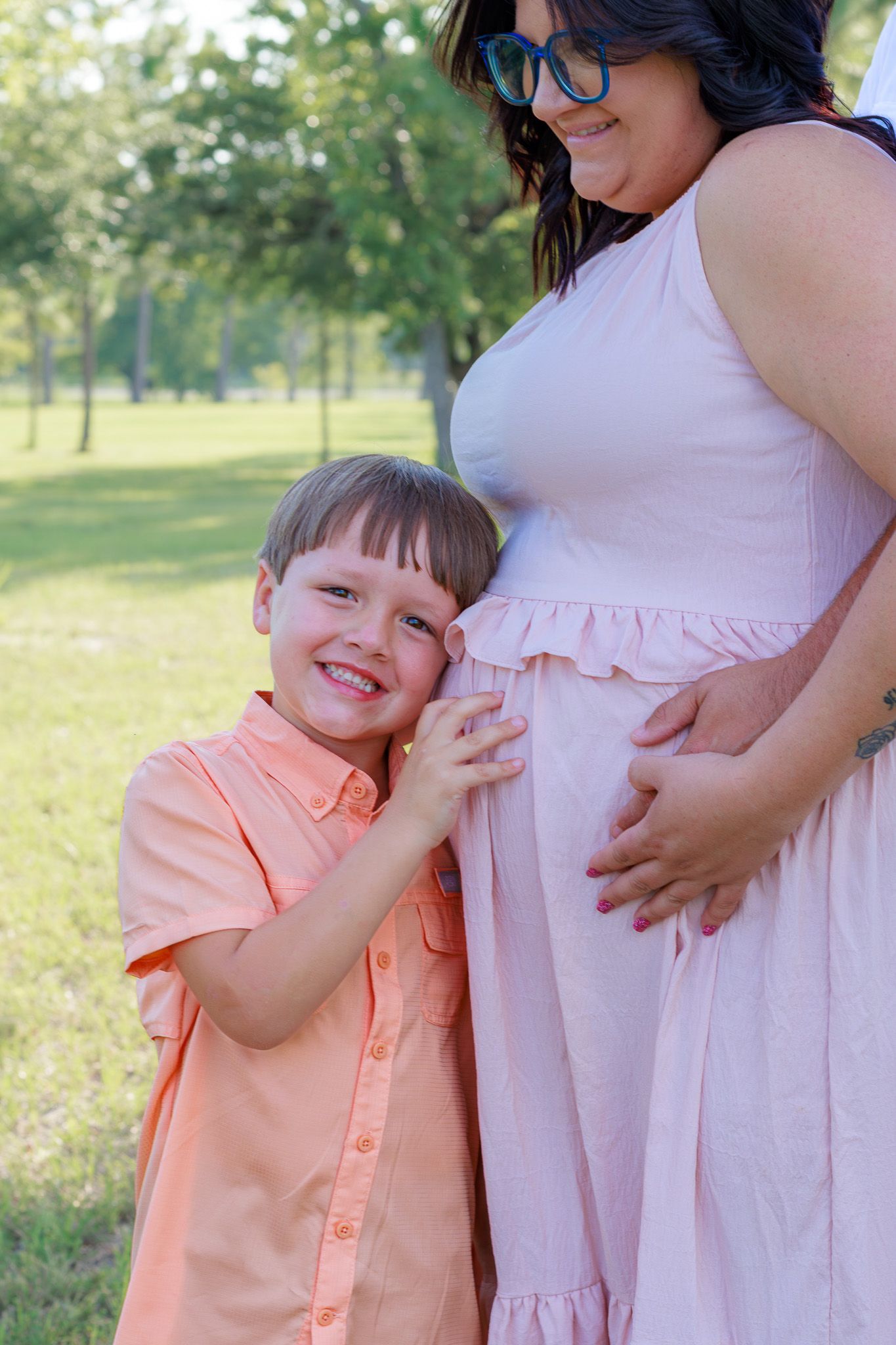 Smiling young boy listens to pregnant mothers belly during an outdoor maternity photo session.