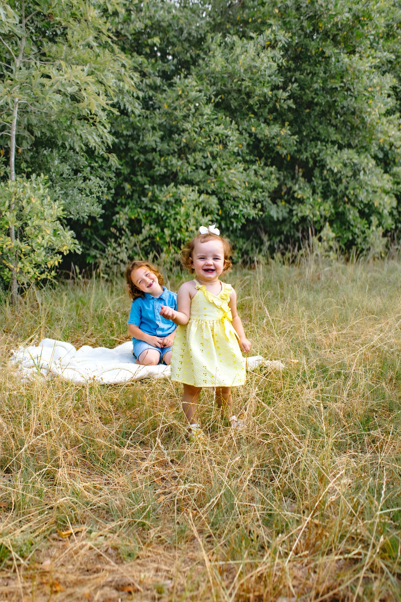 Two young children laughing and playing on a white blanket in a sunny grassy meadow.