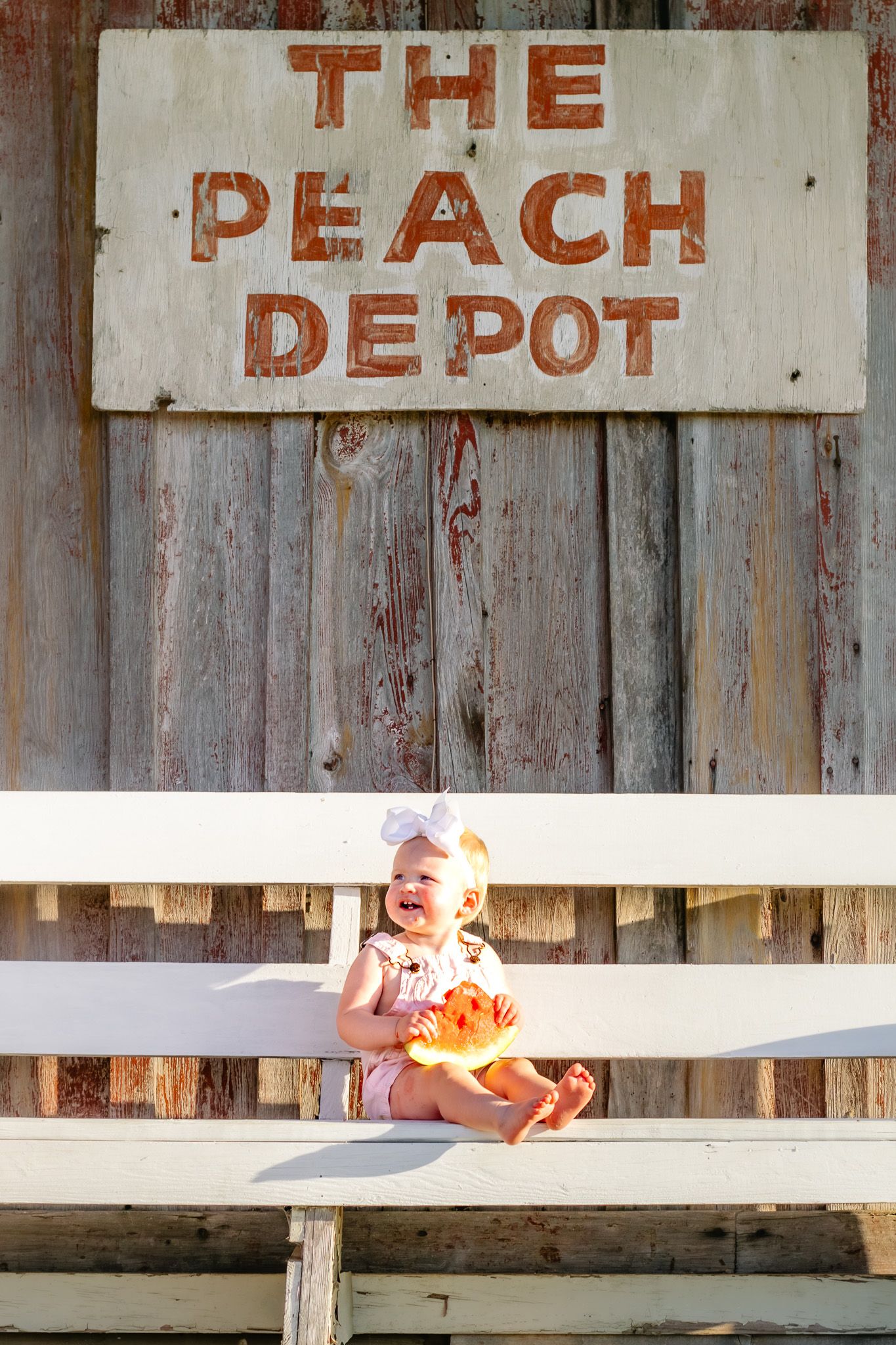Happy baby girl sitting on white bench below rustic Peach Depot sign, holding a peach outdoors.