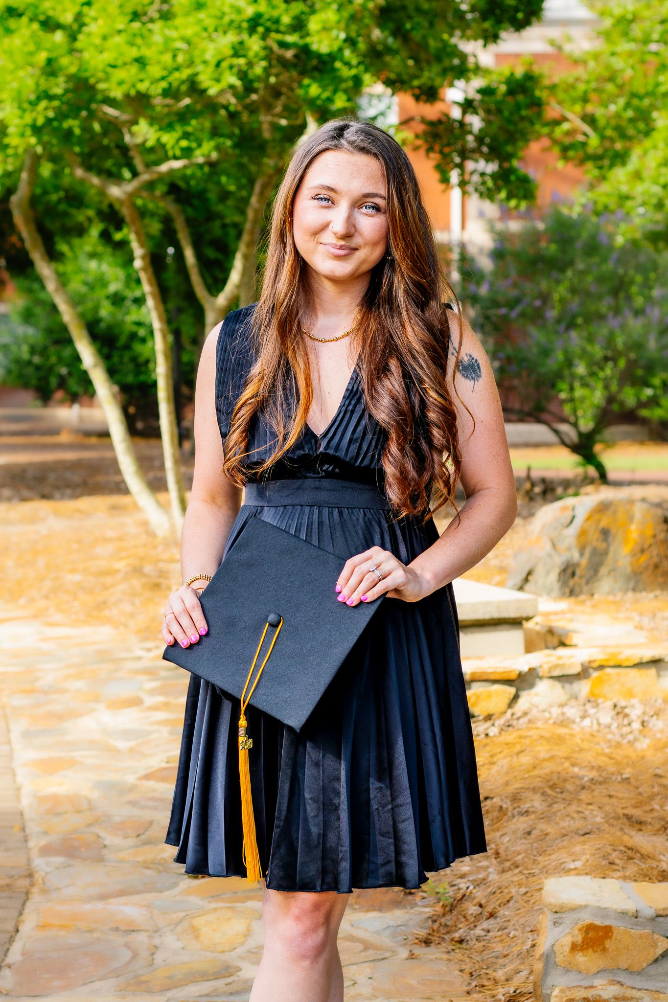 Young woman in navy dress holds black graduation cap with gold tassel outdoors in sunny setting.