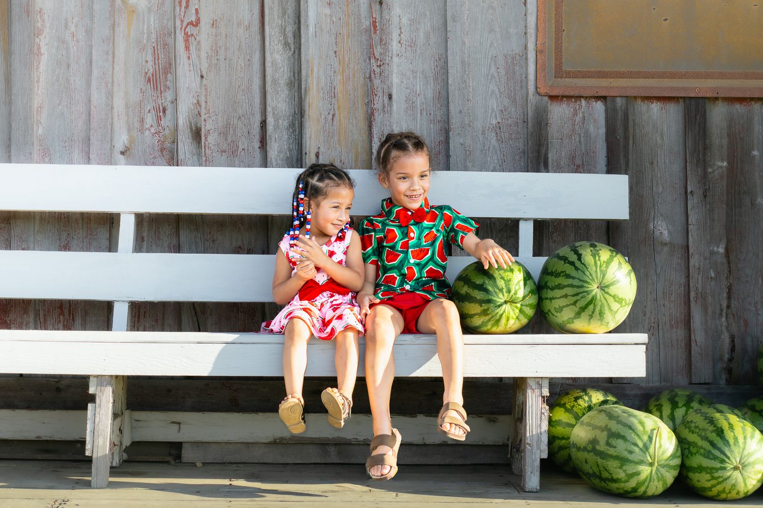 Two children sit on white bench with watermelons at a farm, wearing colorful summer outfits.