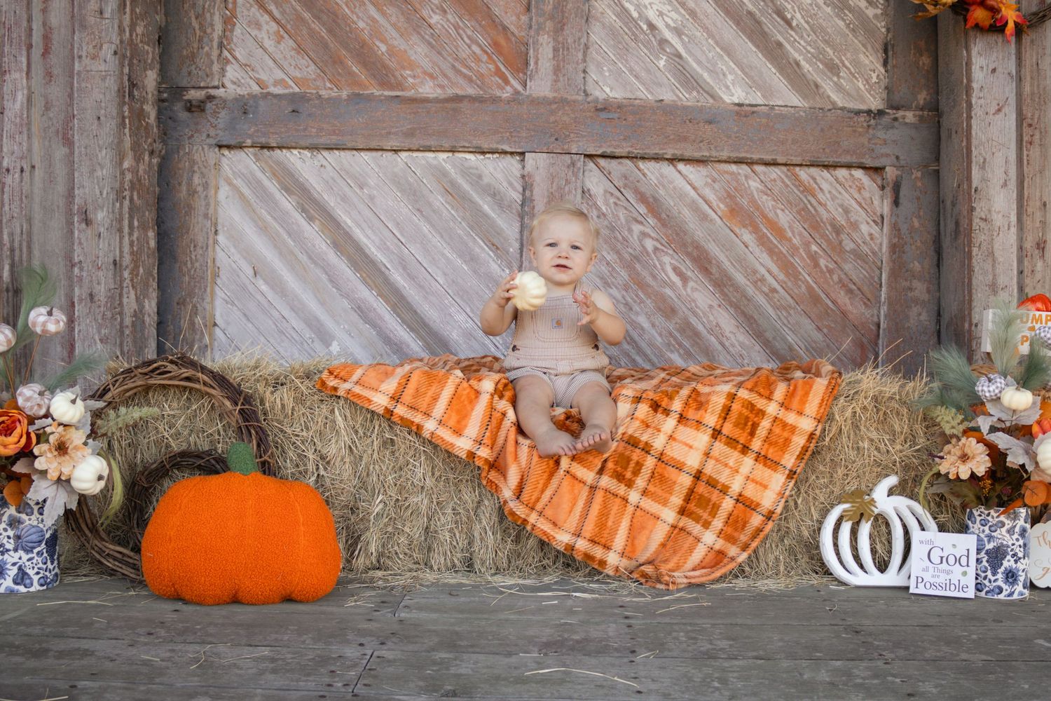 Baby sitting on hay bale with orange plaid blanket and pumpkins in fall barn photo session.