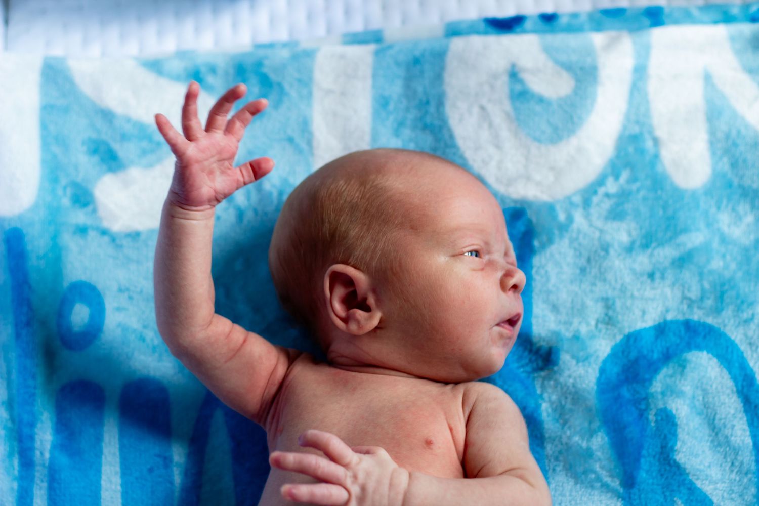 Newborn baby lying on blue blanket with arm raised and hand outstretched.