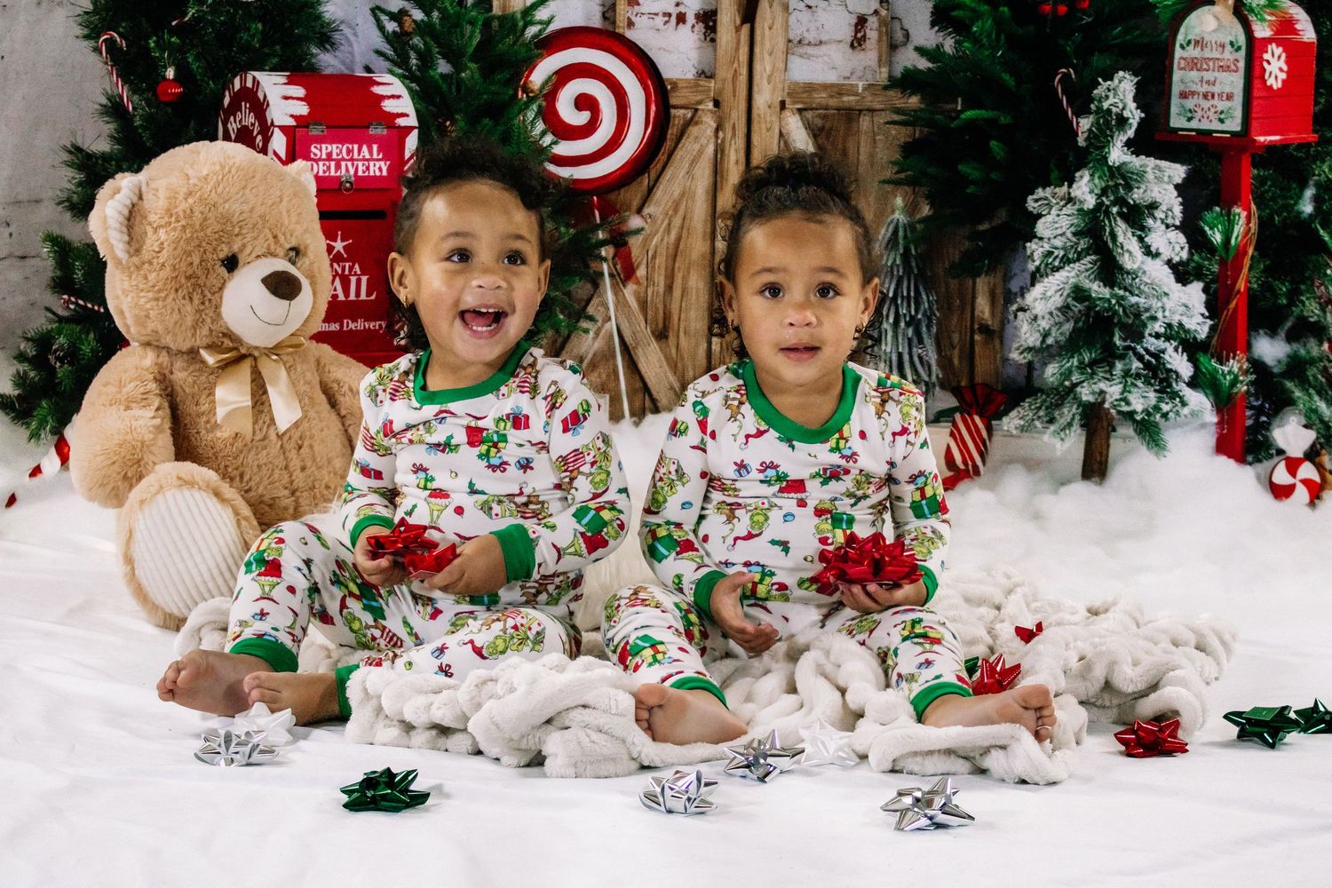 Two young toddler boys in Christmas pajamas sitting with teddy bear and holiday decorations.