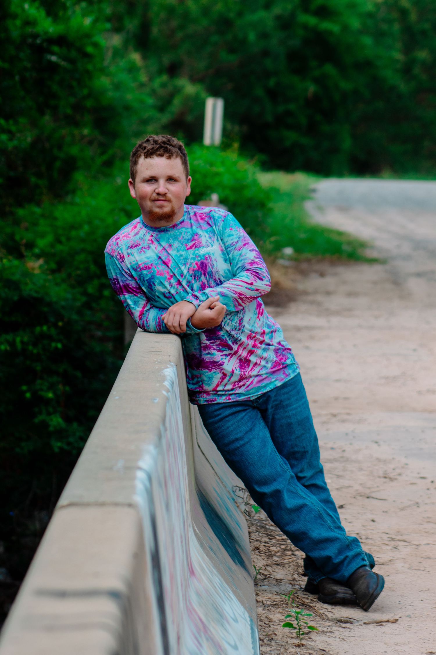Young man in tie-dye shirt posing against a bridge railing on a rural road.