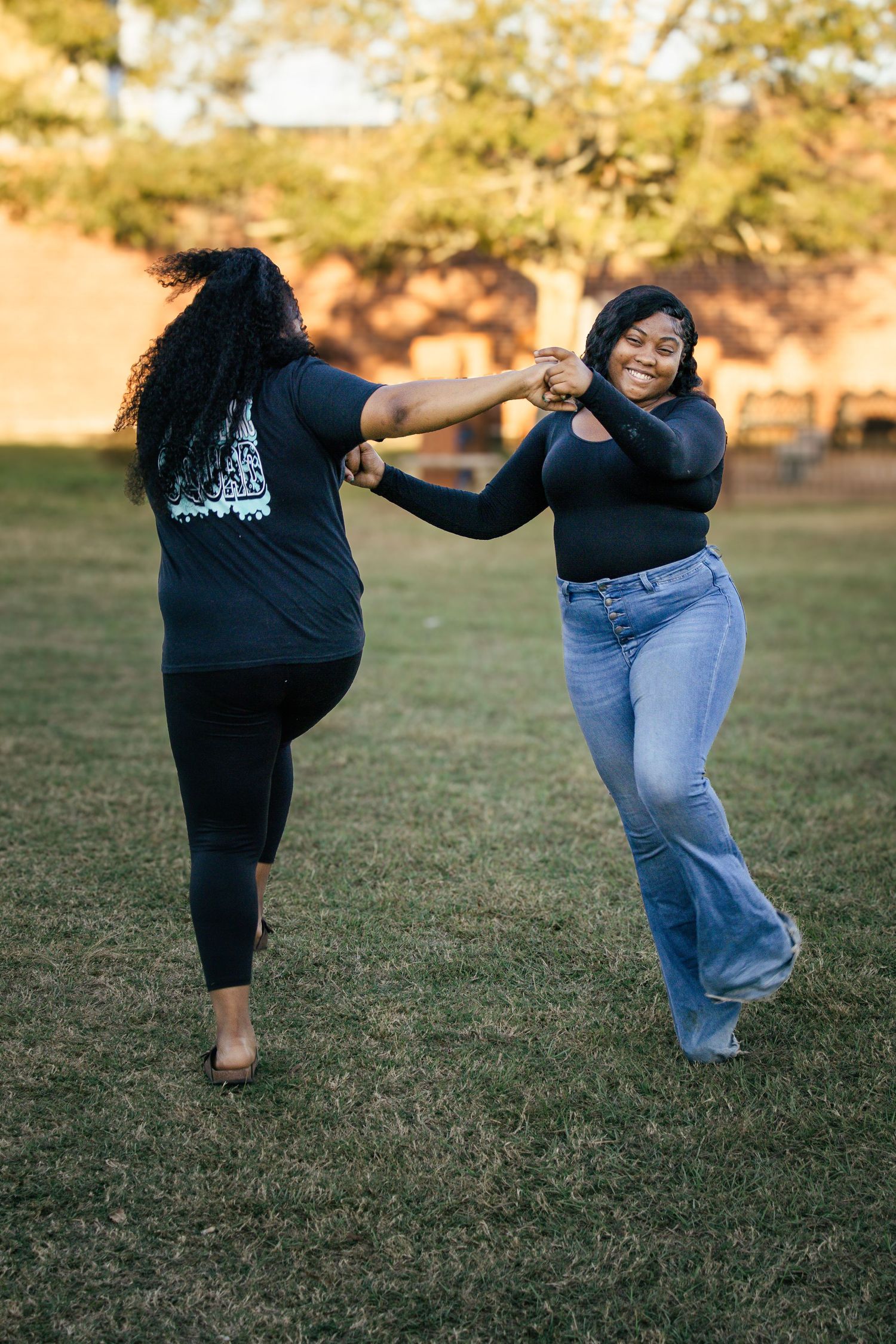 Two young Black women laughing and spinning together holding hands on a sunny green lawn.