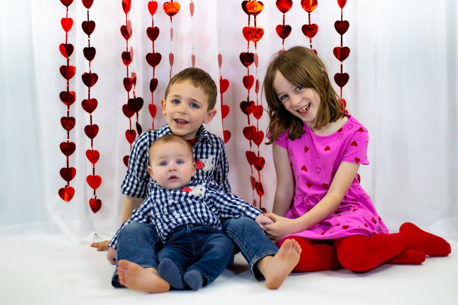 Three siblings pose together in Valentine's Day outfits before a backdrop of hanging red heart garlands.