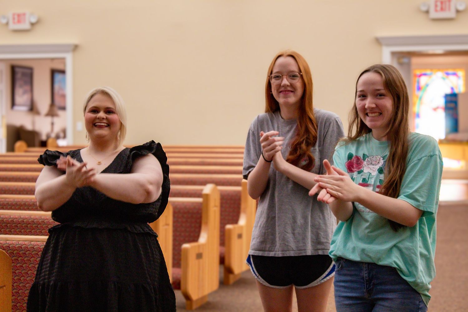 Three smiling women stand in a church sanctuary with wooden pews clapping and holding a phone.