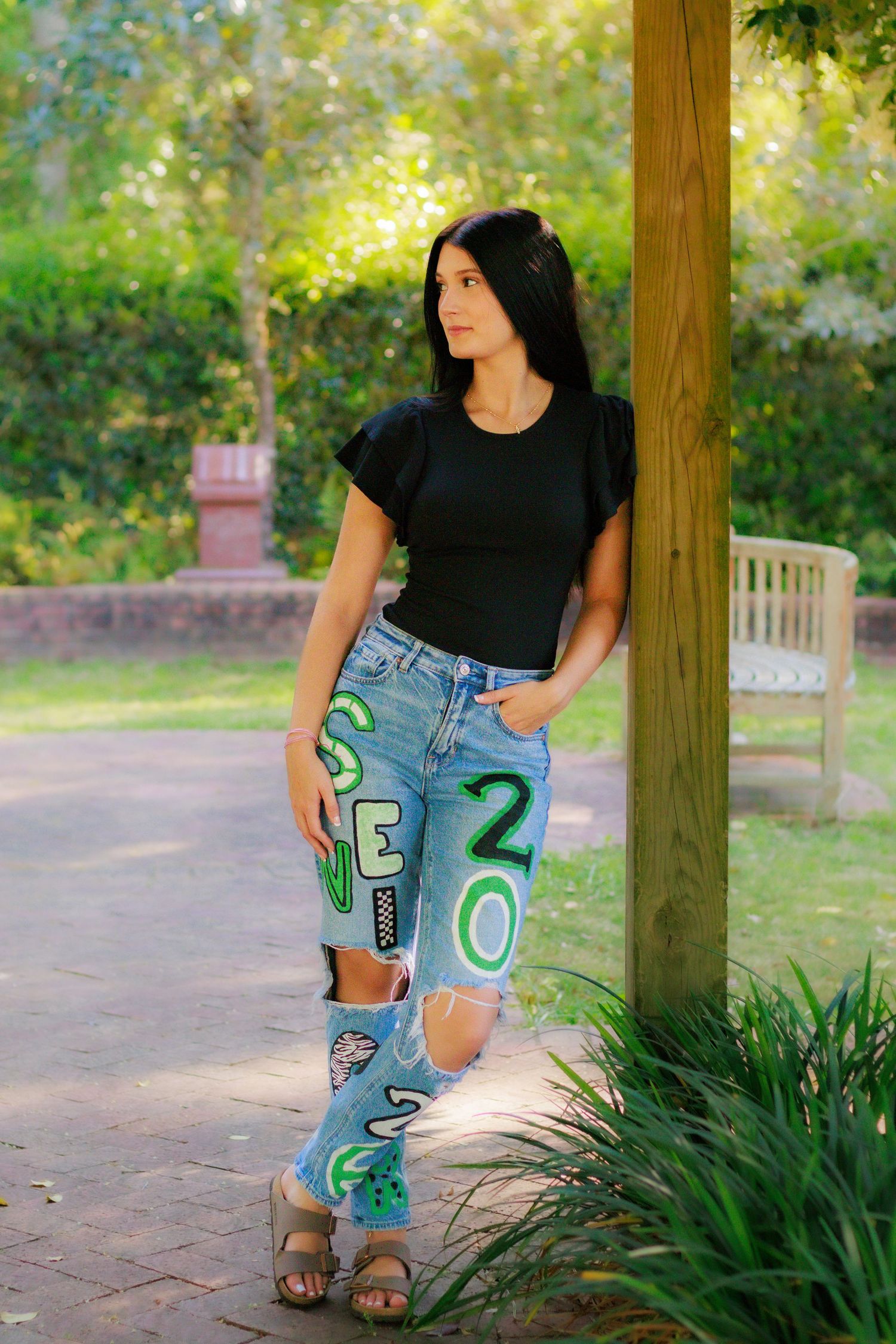 Young woman in black top and custom painted graduation jeans poses by a tree in a sunny park.