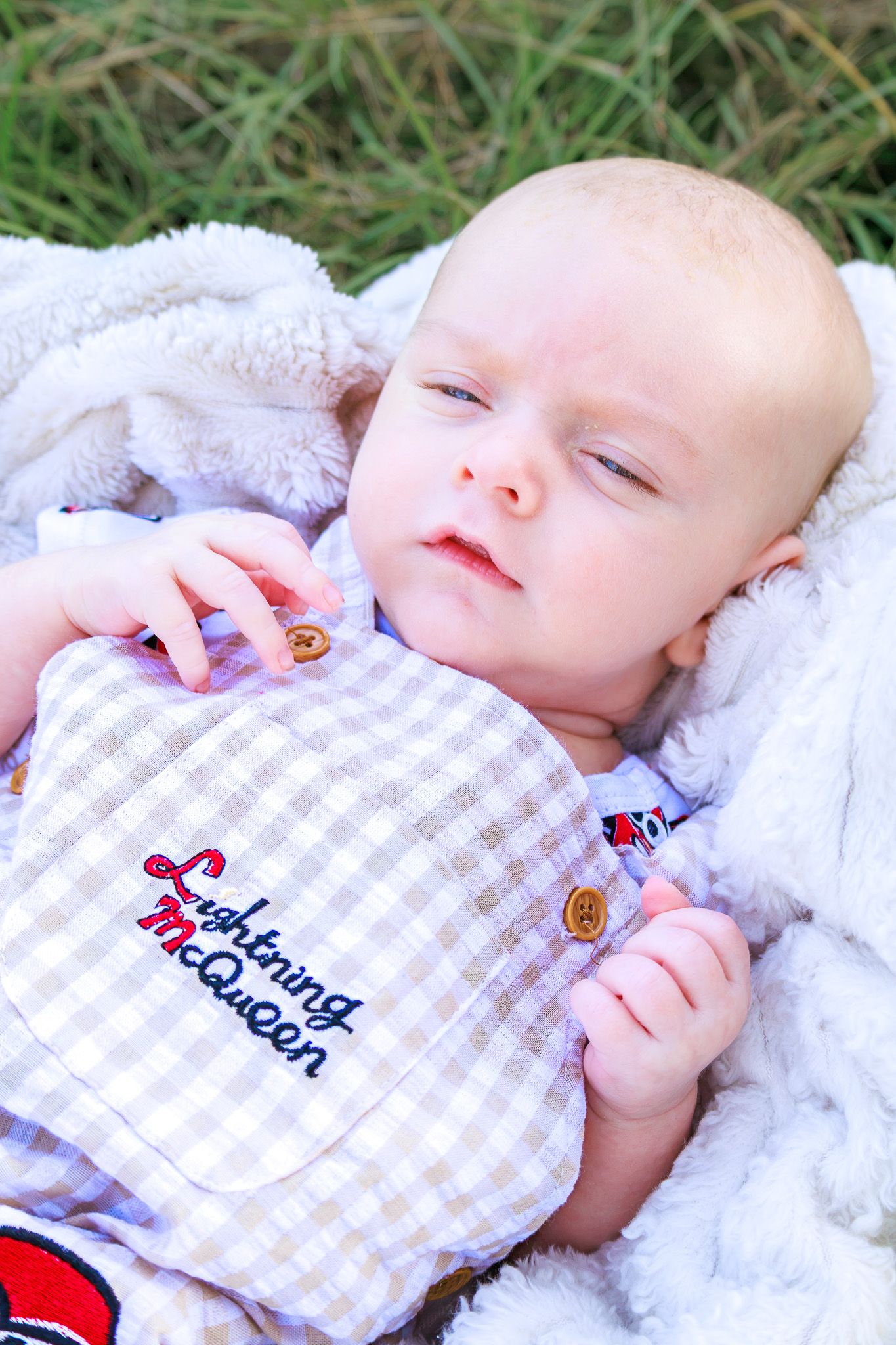 Baby wearing a Lightning McQueen gingham outfit lying on a white fluffy blanket outdoors on green grass.