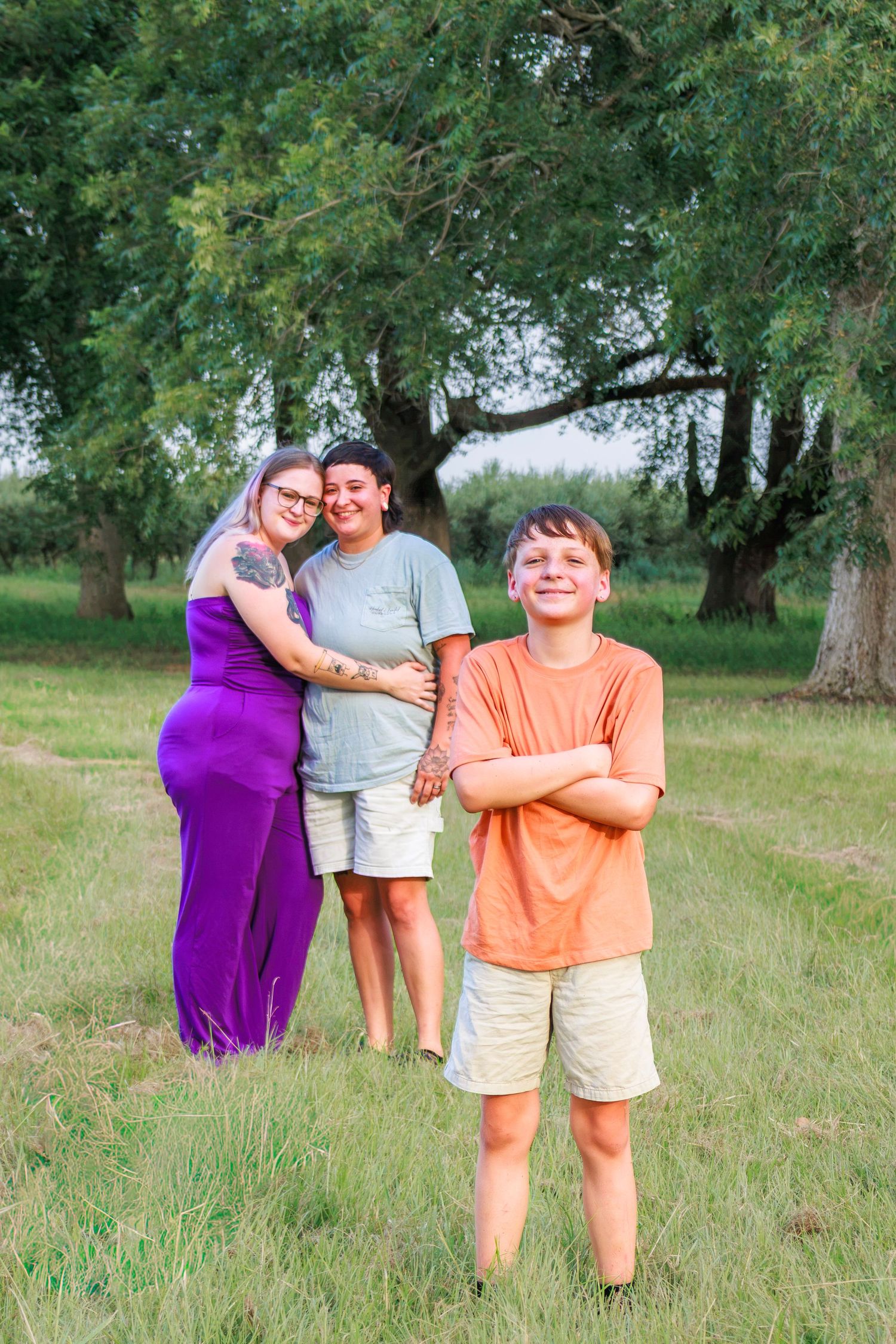 A smiling boy in an orange shirt stands with arms crossed in a green field, while two women embrace warmly behind him.