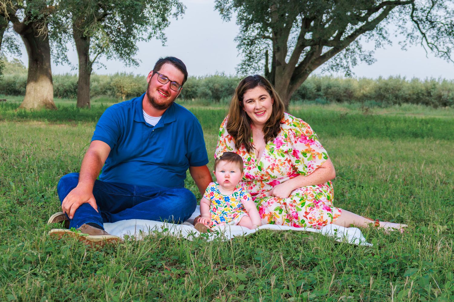 Family portrait of parents and baby sitting on blanket in green park under trees.