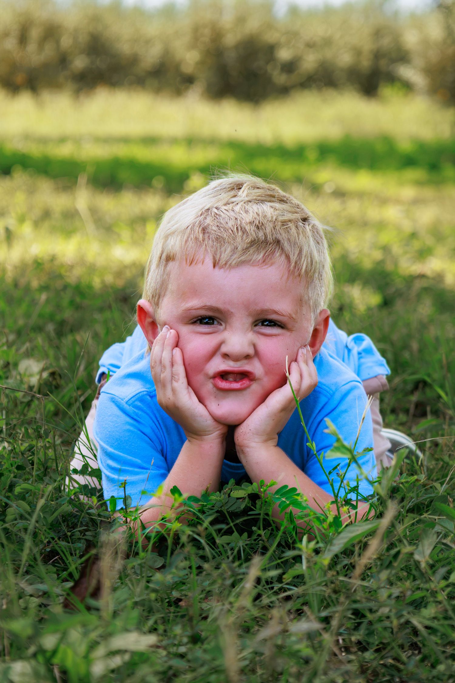 Blonde boy lying on grass with hands on cheeks, making a funny face outdoors in a green field.
