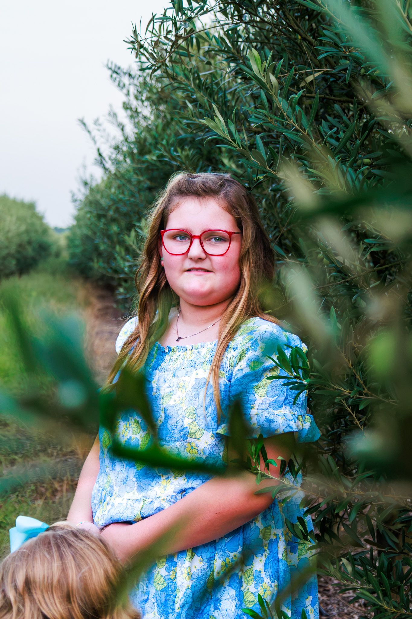 Young girl with red glasses in a blue floral dress standing among green olive trees outdoors.