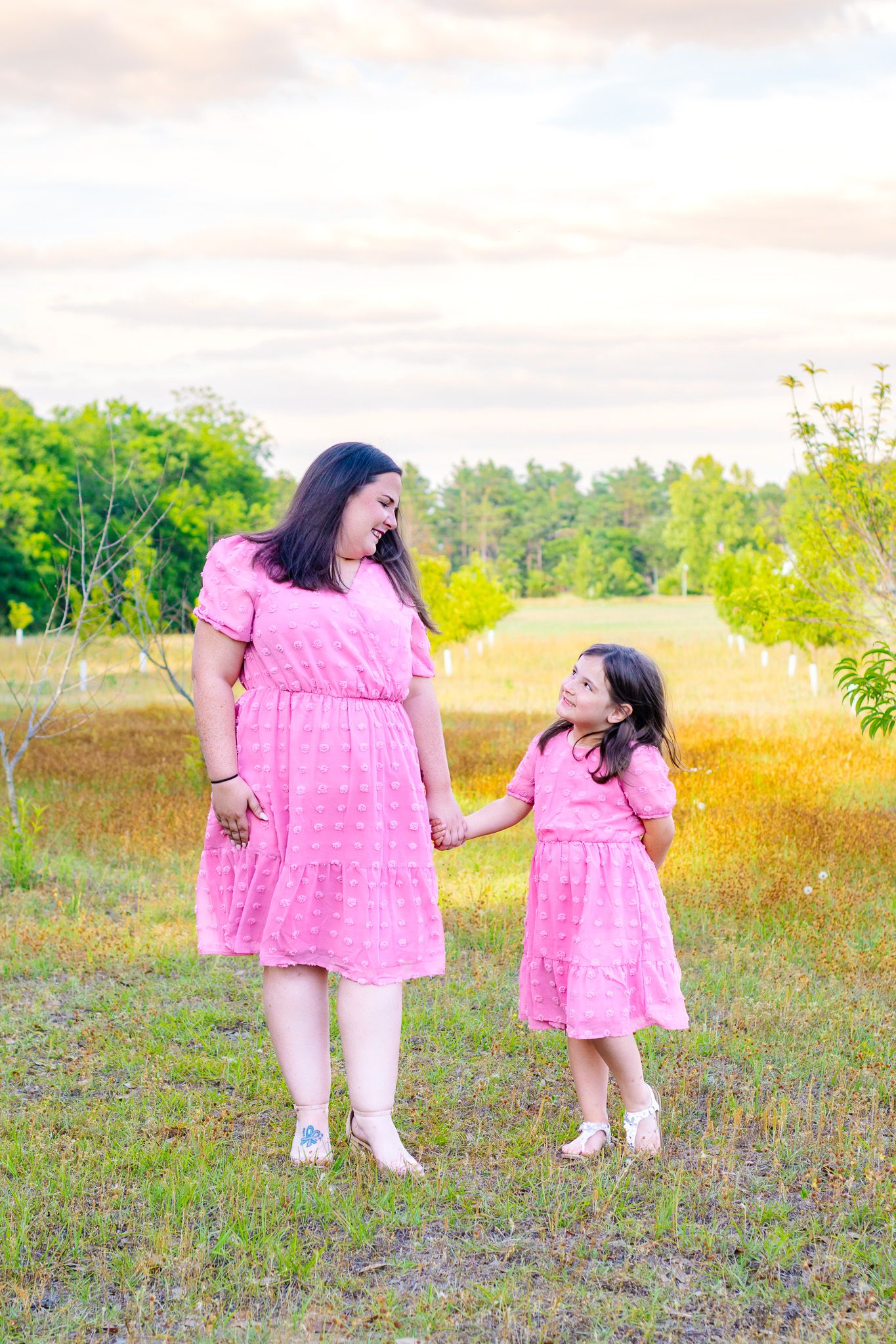 Mother and daughter in matching pink dresses holding hands in a sunny field.