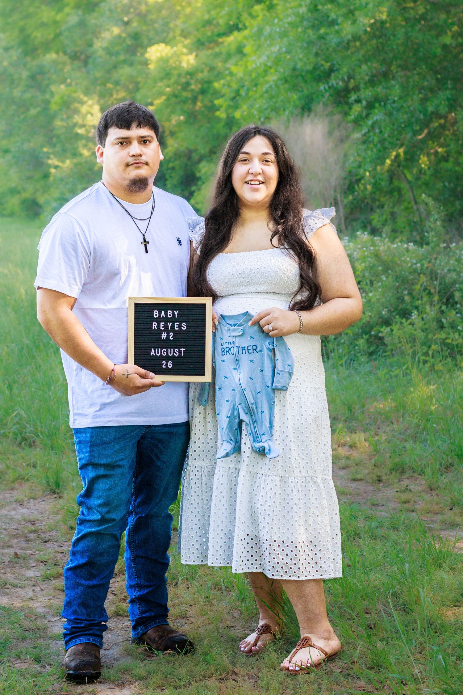 Couple stands outdoors holding a letter board announcing Baby Reyes #2 and a blue baby outfit.