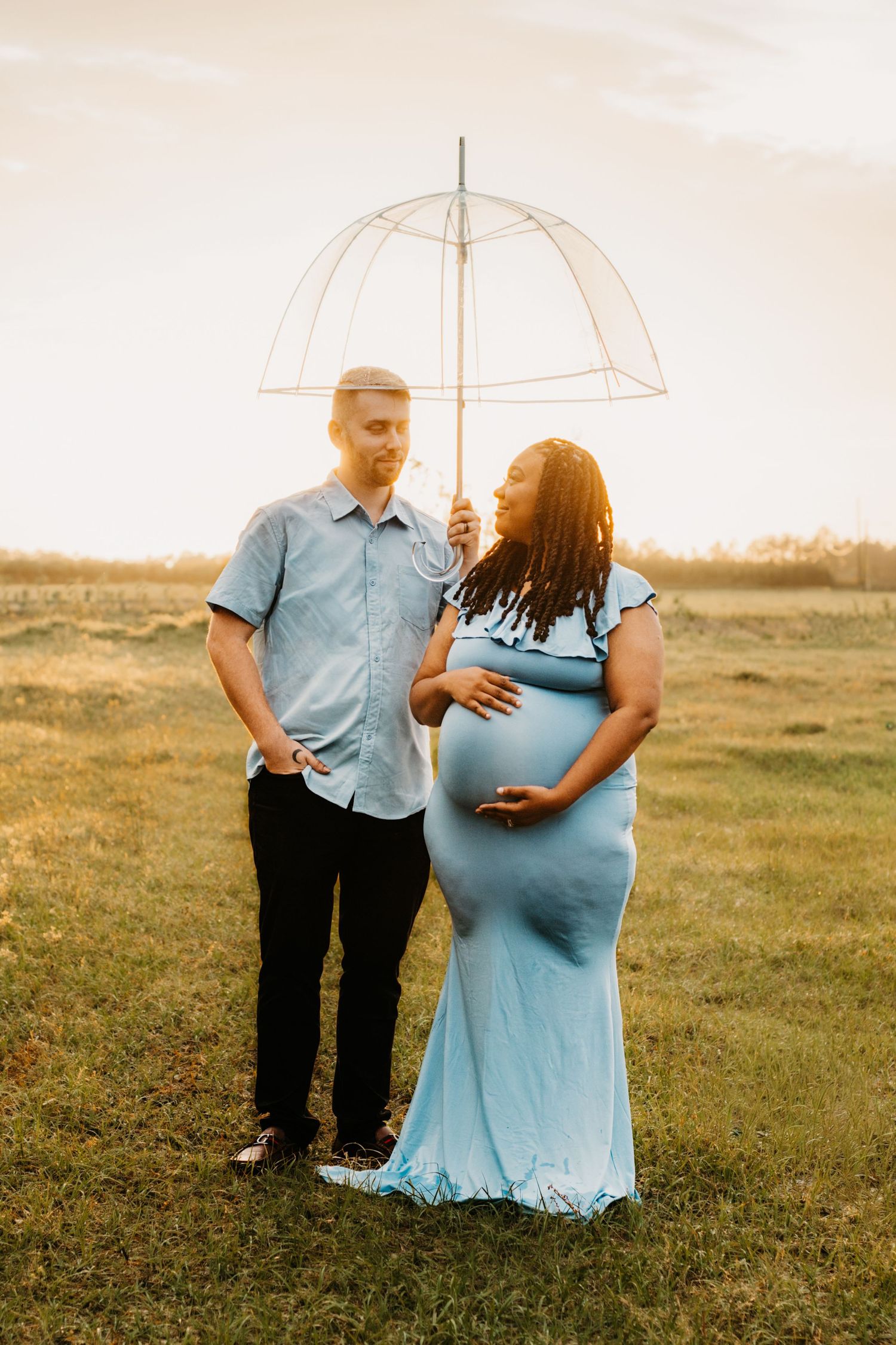 Interracial couple poses in golden sunset field, pregnant woman in blue gown holding clear umbrella.