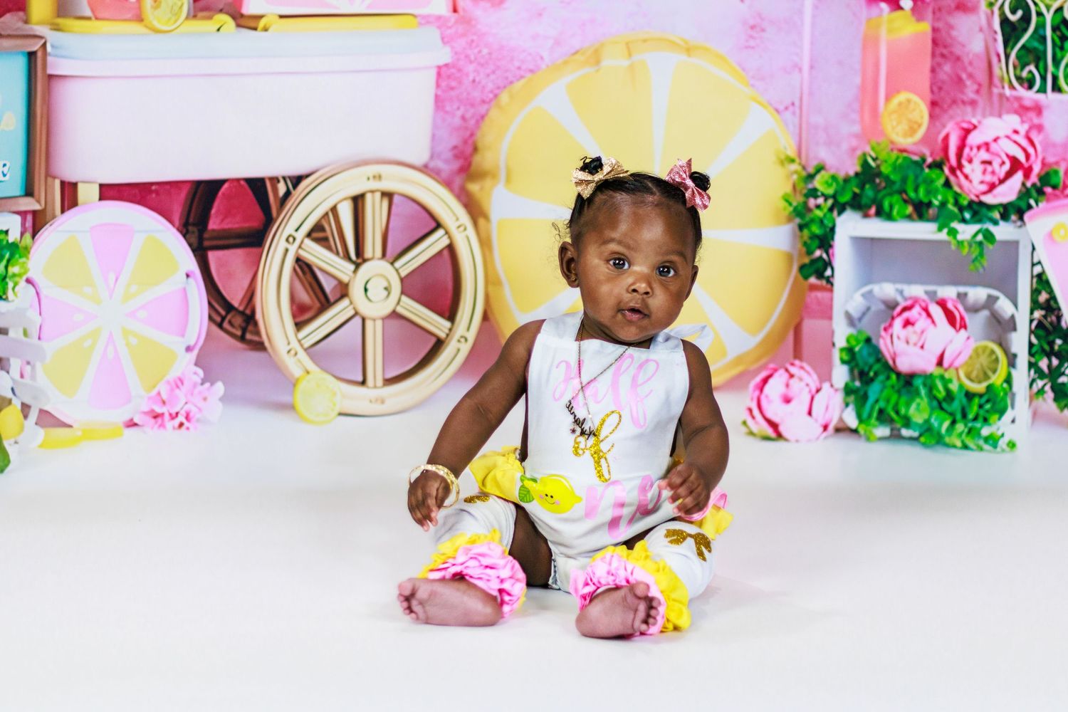 Adorable baby girl in a pink and yellow lemon outfit sitting in front of a colorful lemonade stand backdrop.